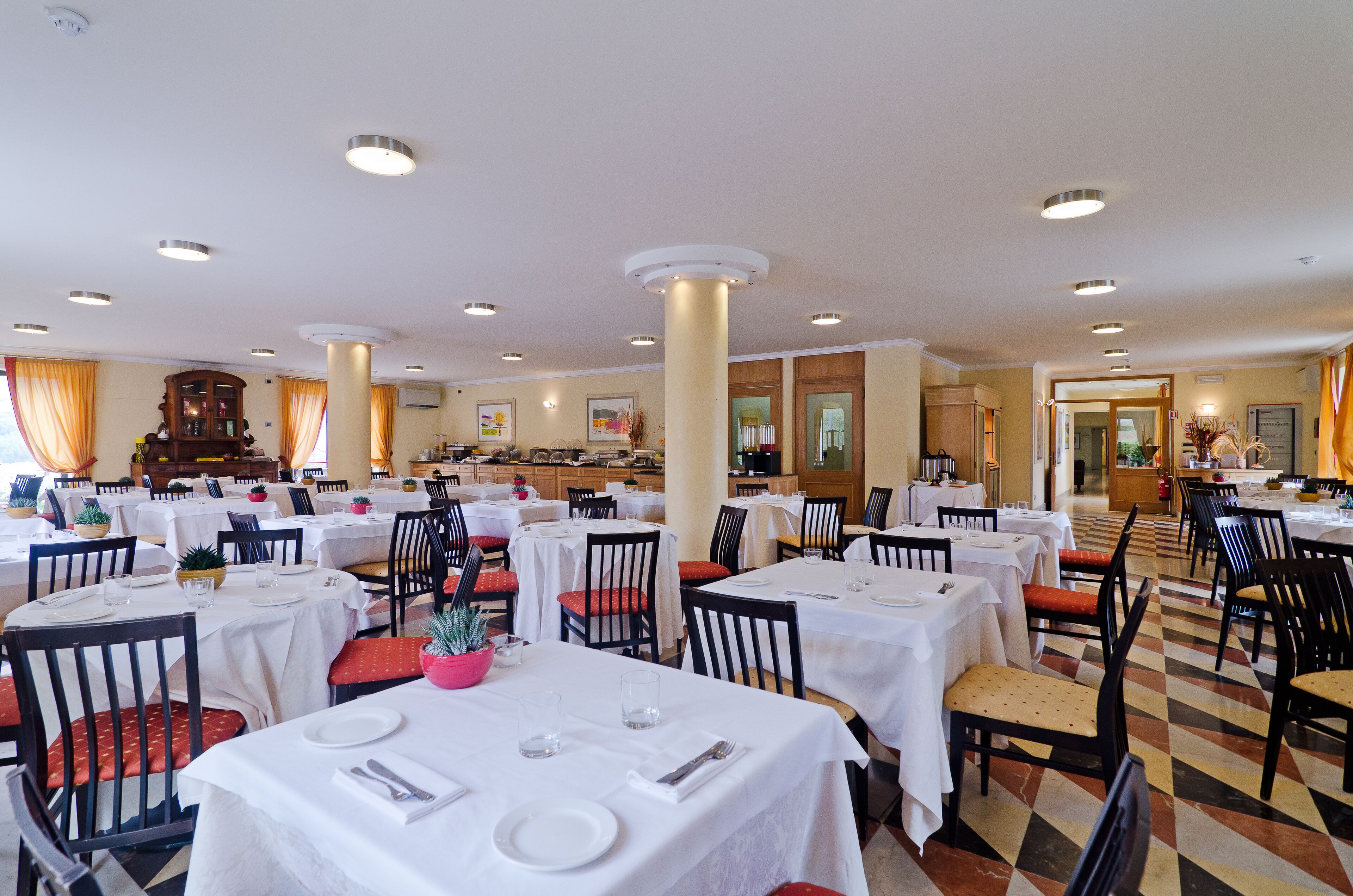 Un vaste espace dédié à la salle à manger, avec de nombreuses tables et chaises, une colonne centrale et un motif de carreaux au sol.