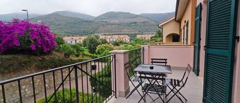 A balcony with a table and chairs, overlooking a hillside with buildings and lush greenery.