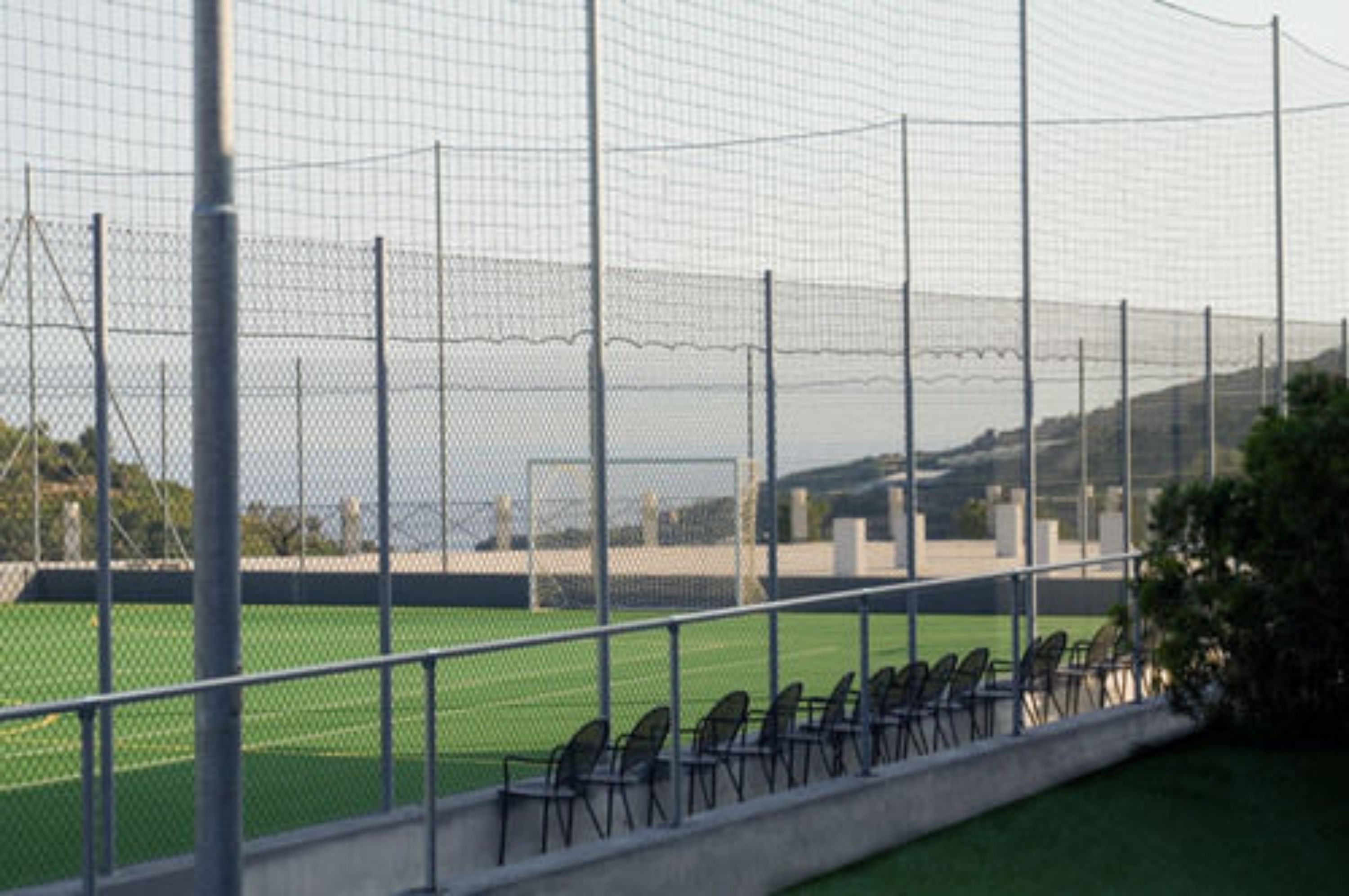 Un campo deportivo con una estructura de malla, sillas a un lado y una vista despejada del paisaje circundante.