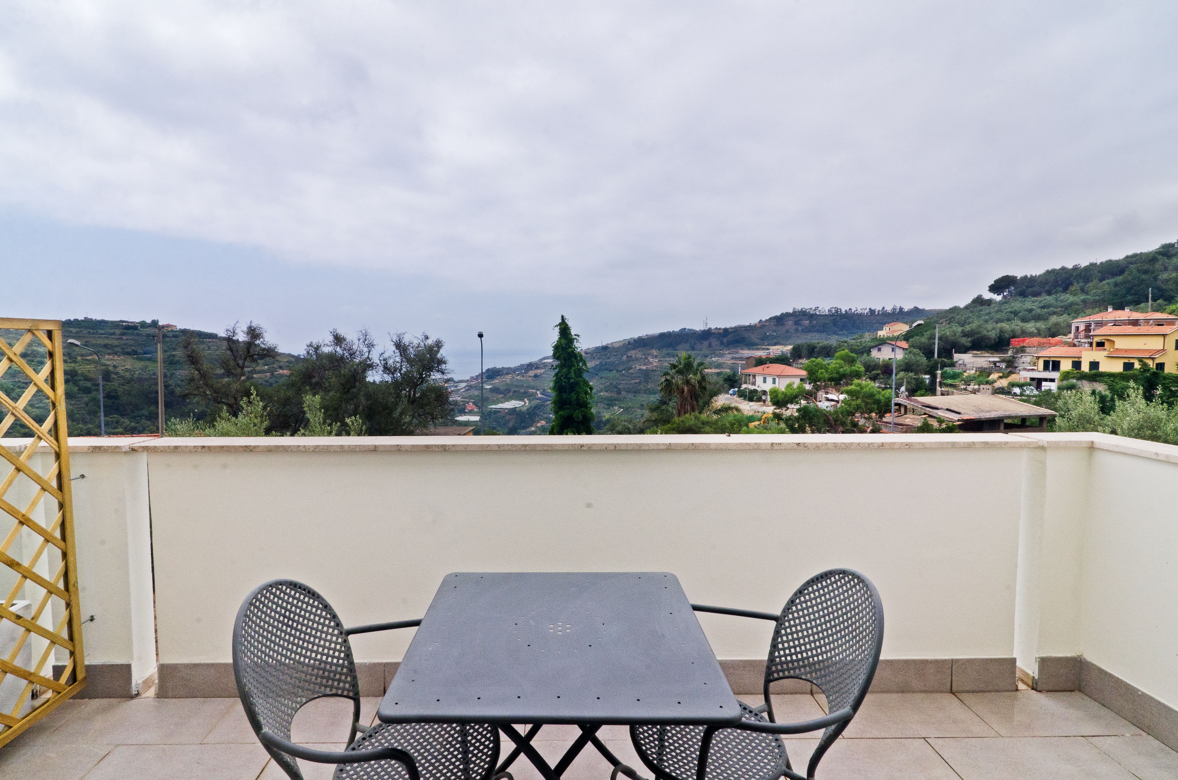 Un balcon avec une table et deux chaises, donnant sur une colline parsemée de bâtiments et de verdure.