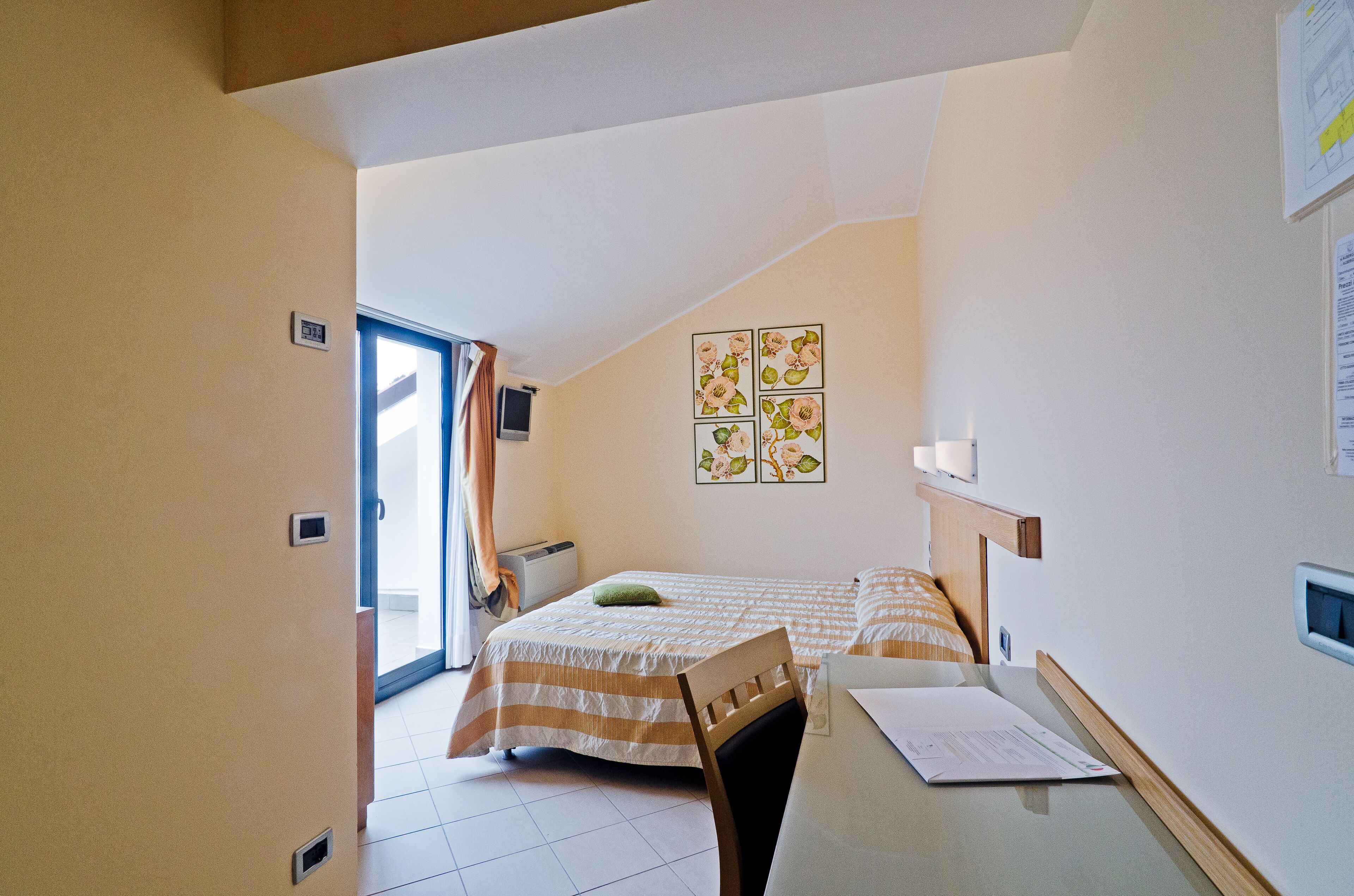 A bedroom with a bed, a desk, and a chair. There is a window with curtains, a wall-mounted TV, and a door leading to another room.