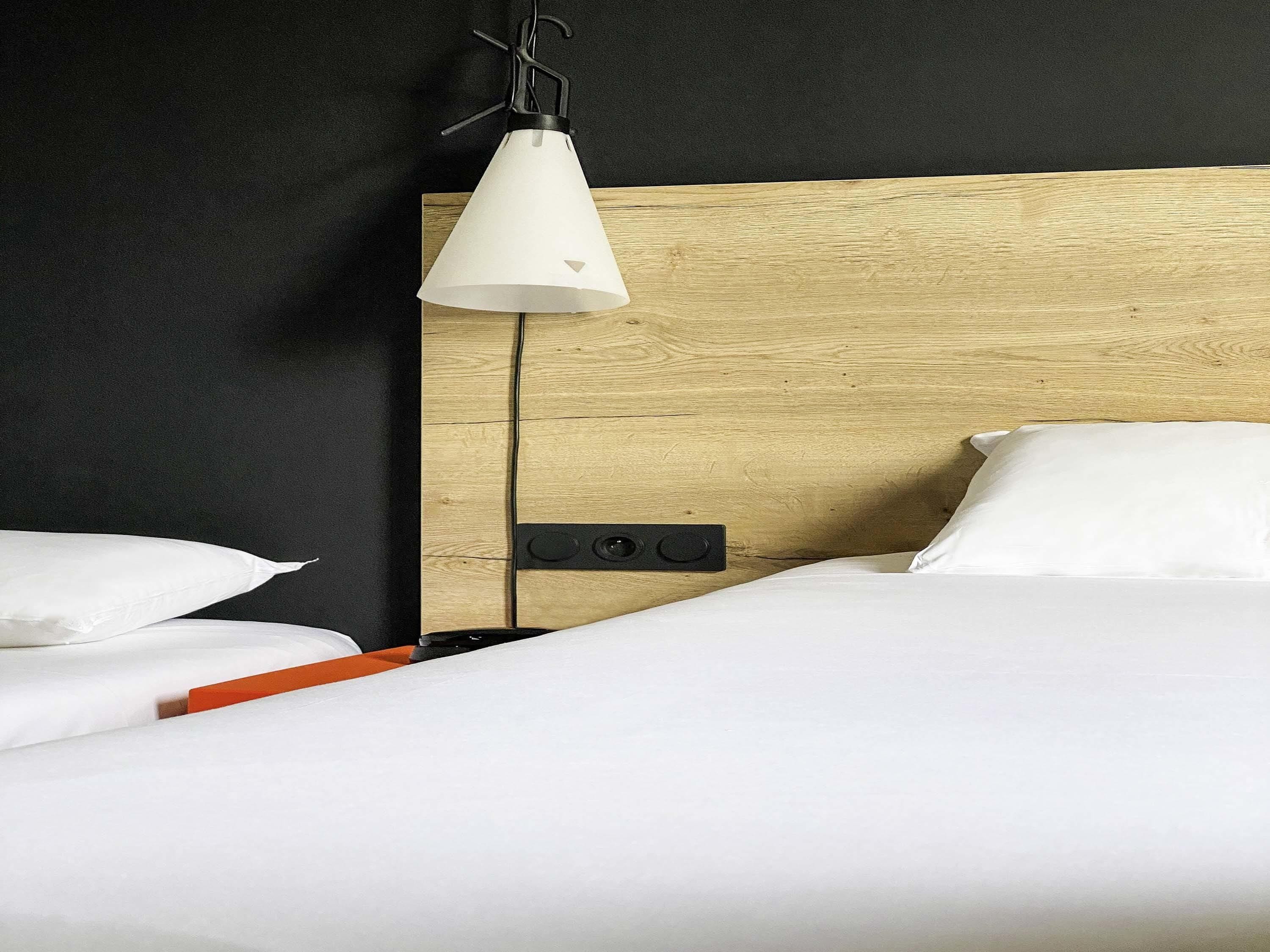 Habitación de hotel con cabecera de madera, ropa de cama blanca, mesita de noche con un objeto rojo y una lámpara colgante.