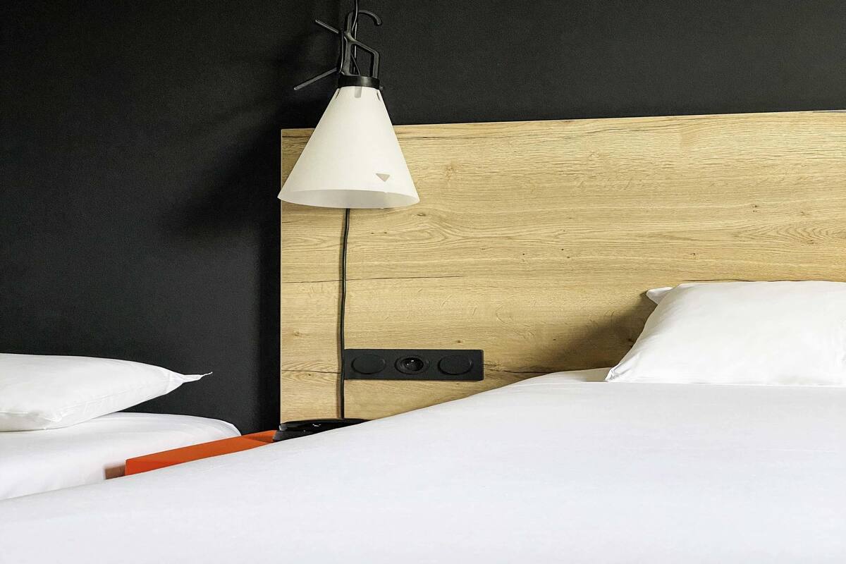 A hotel room with a wooden headboard, white bedding, a bedside table with a red object, and a hanging lamp.