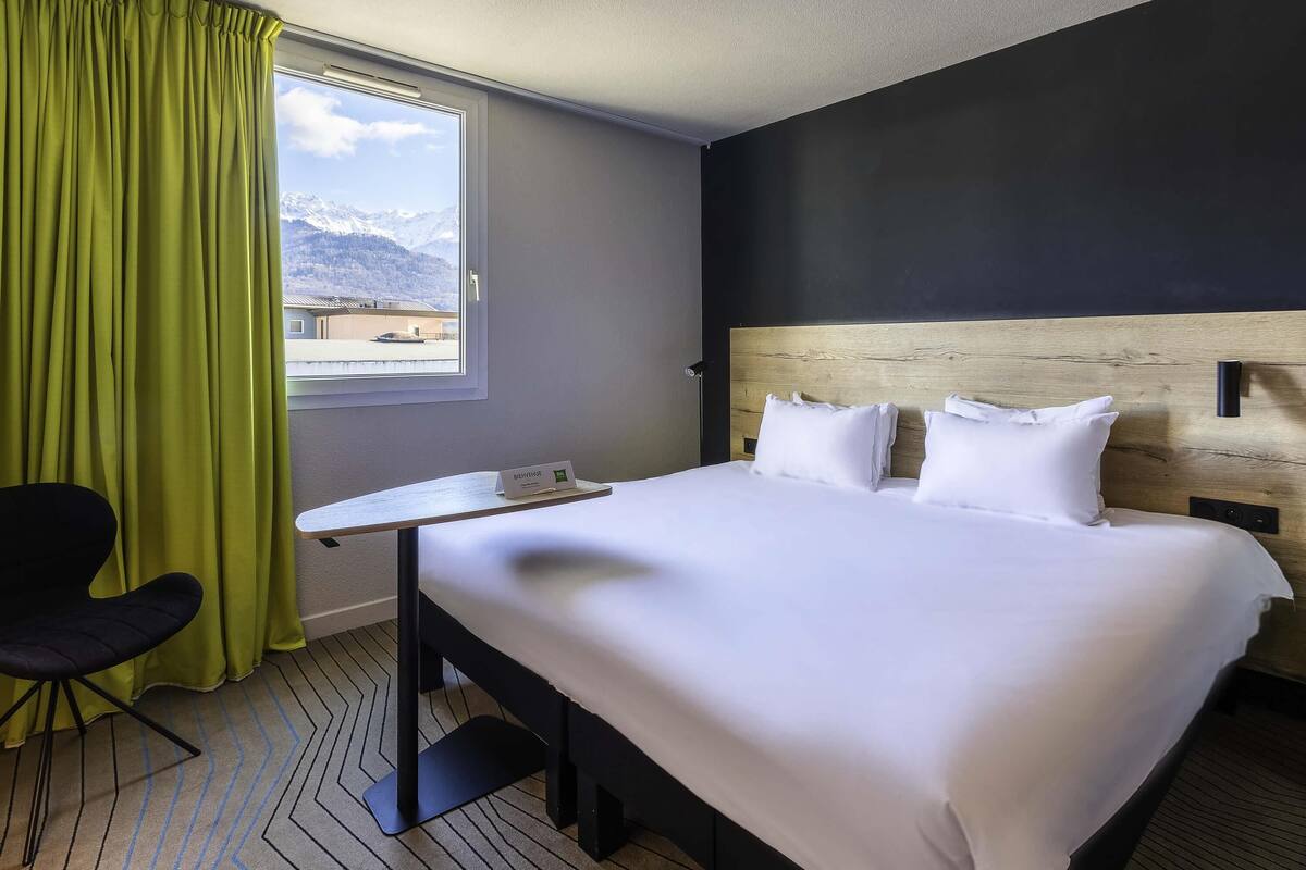 A hotel room with a bed, a desk, a chair, a window with a view of mountains, and a nameplate on the desk.