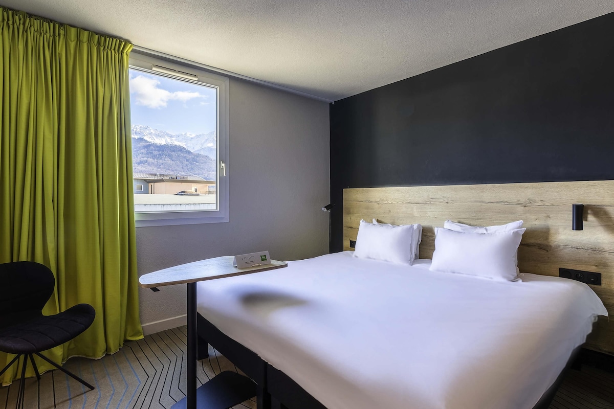 Habitación de hotel con cama, escritorio, silla, ventana y vistas a montañas, además de una placa con el nombre sobre el escritorio.