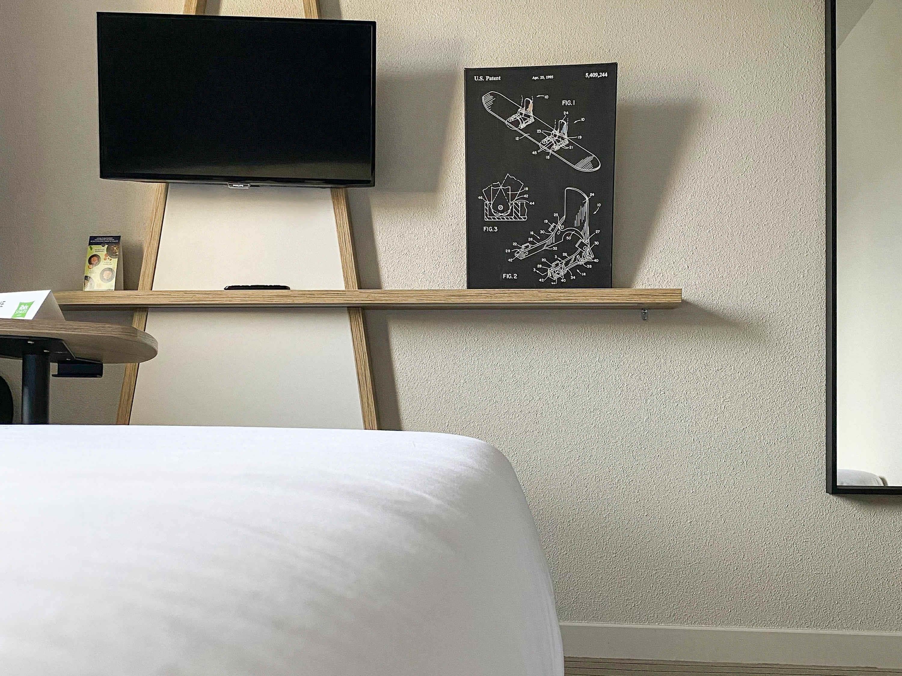 Una habitación de hotel con cama, televisor montado en la pared, estante con un dibujo de patente y una pequeña mesa con un letrero.
