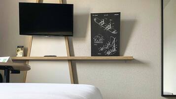 Una habitación de hotel con cama, televisor montado en la pared, estante con un dibujo de patente y una pequeña mesa con un letrero.