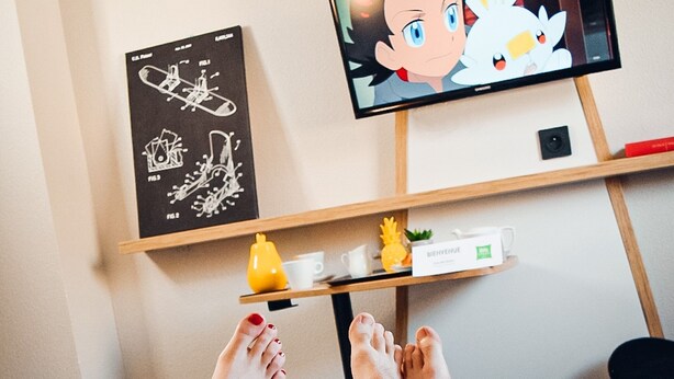 A person lying on a bed with feet up, holding a remote control, and a television displaying an animated character. - ibis Styles Crolles Grenoble A41 (Crolles)