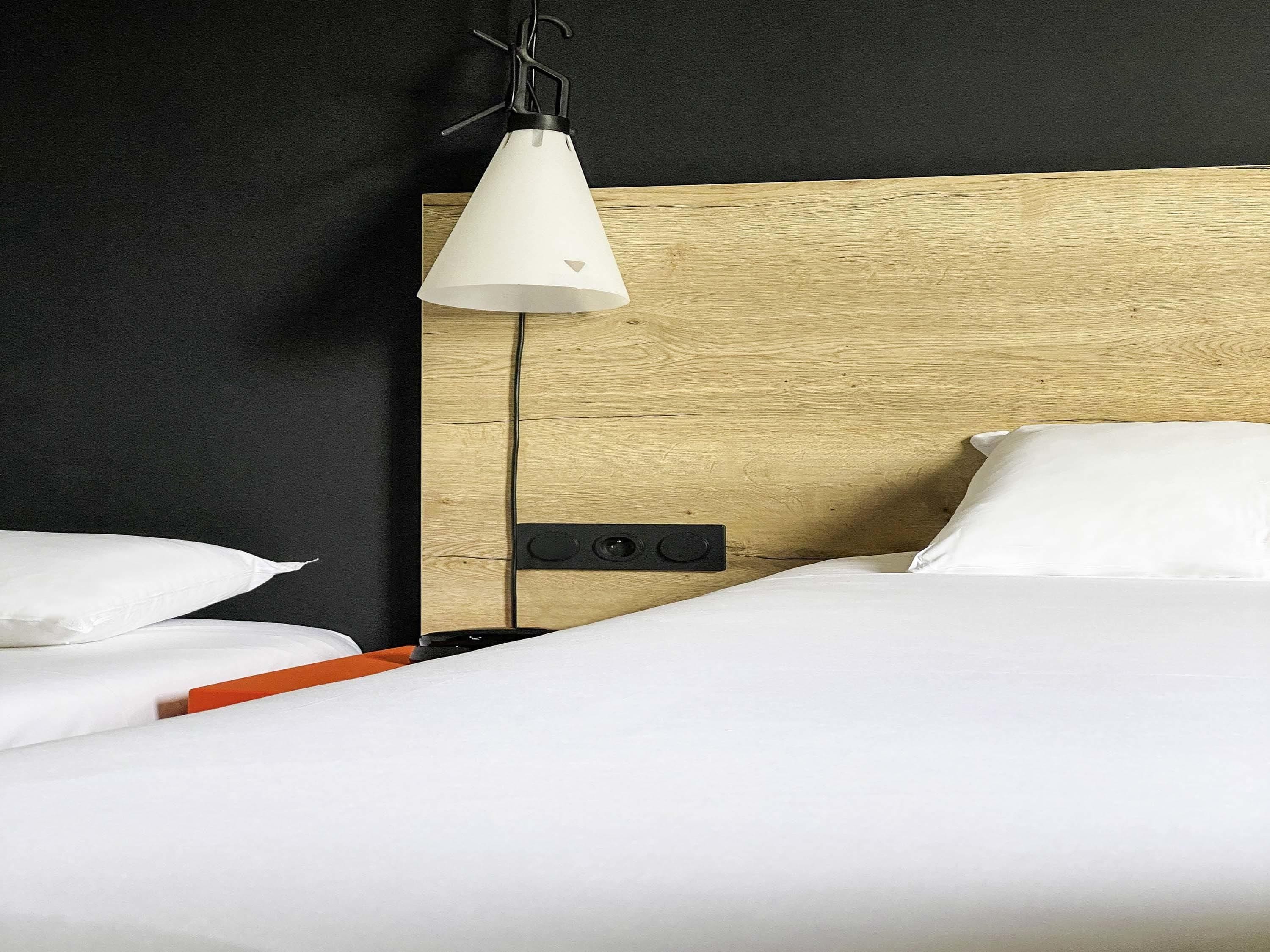 Habitación de hotel con cabecera de madera, ropa de cama blanca, mesita de noche con un objeto rojo y una lámpara colgante.