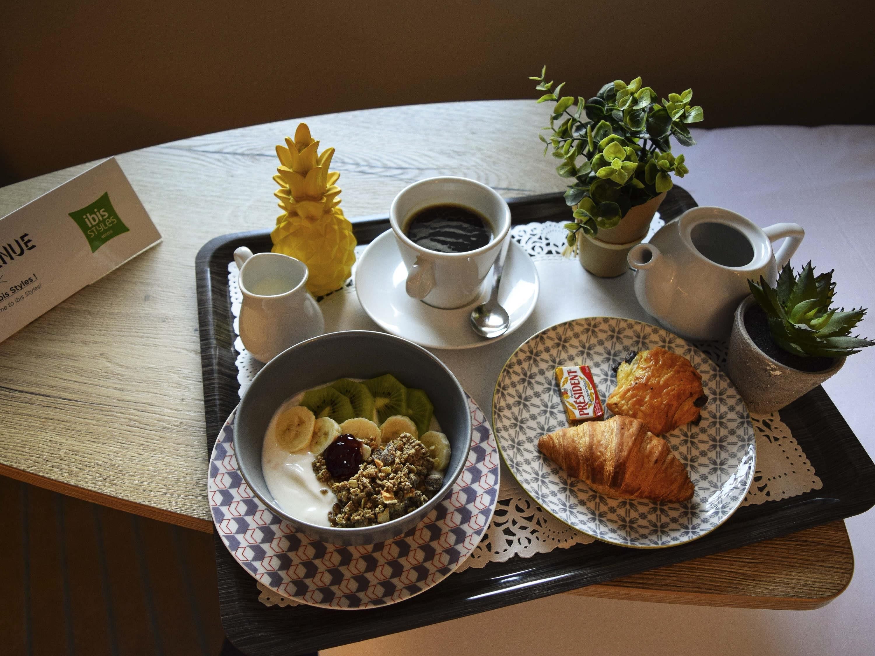 Una bandeja con artículos de desayuno, como un tazón de yogur con frutas, un croissant, un pastel pequeño y dos tazas de café.