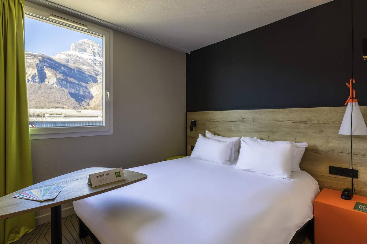 A hotel room with a bed, a table with a welcome brochure, a nightstand with a lamp, and a window with a mountain view.