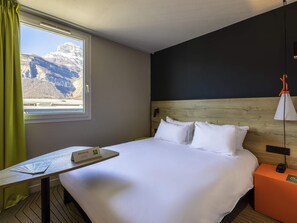 A hotel room with a bed, a table with a welcome brochure, a nightstand with a lamp, and a window with a mountain view. - ibis Styles Crolles Grenoble A41 (Crolles)