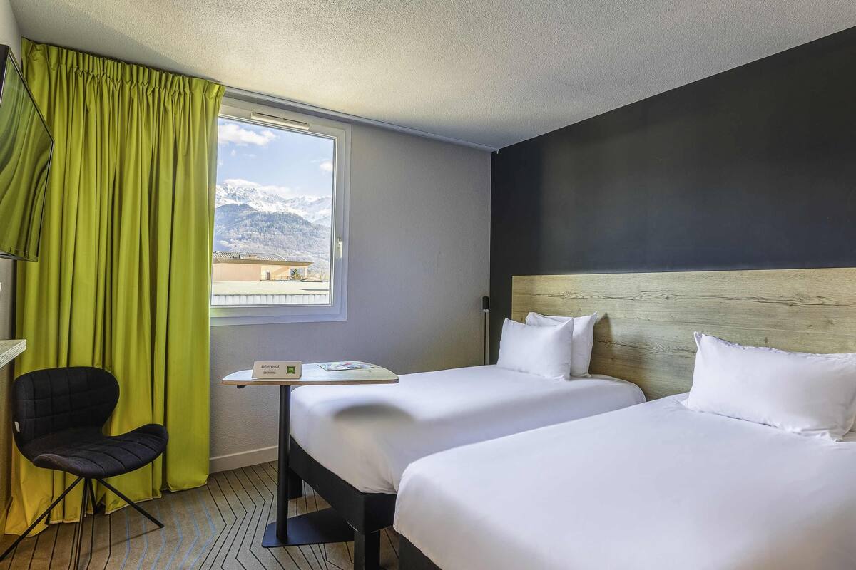 A hotel room with two beds, a desk, a chair, and a window with a view of mountains.