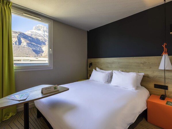 Habitación de hotel con cama, una mesa con un folleto de bienvenida, una mesita de noche con lámpara y una ventana con vista a la montaña.