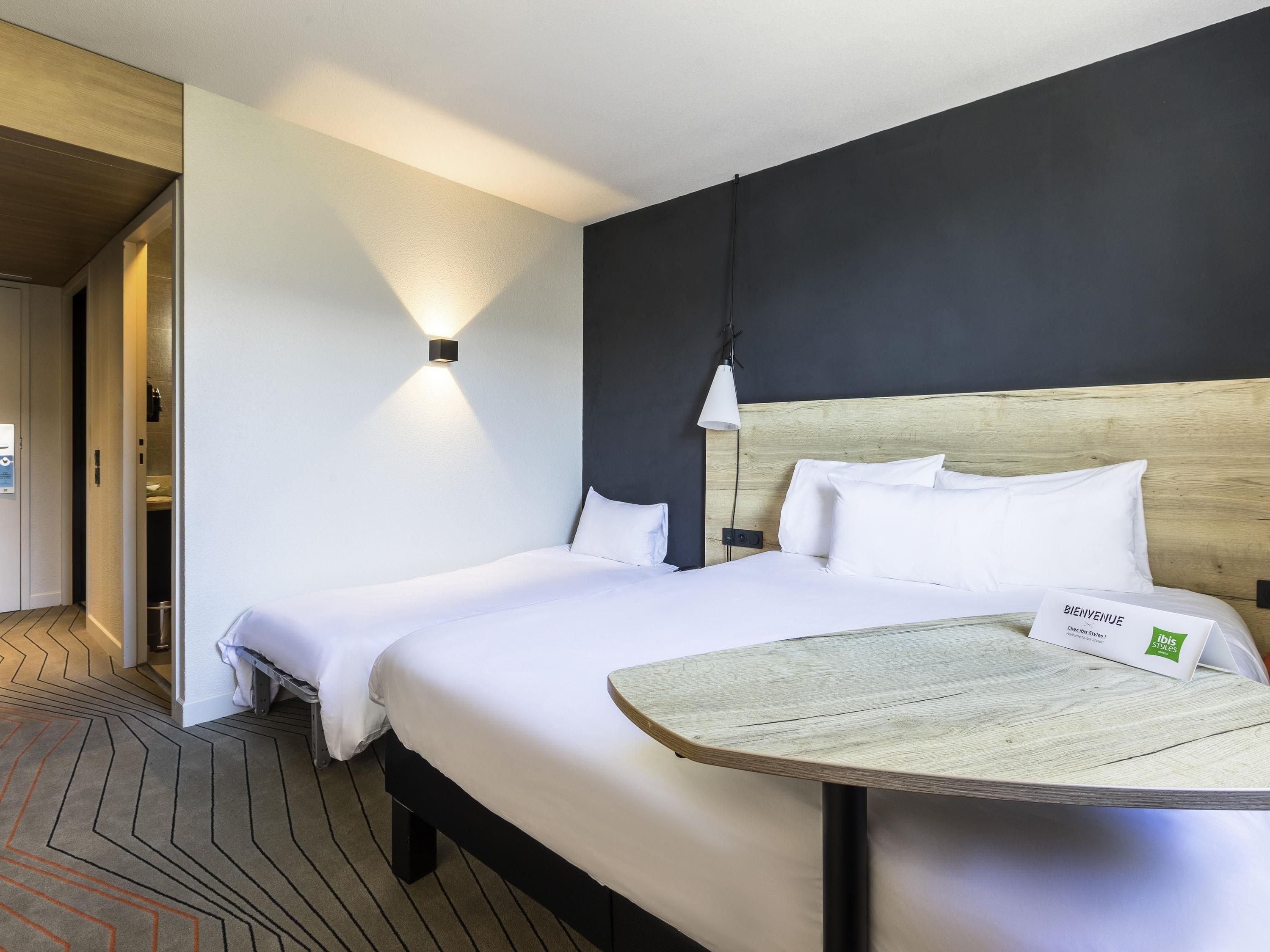 A hotel room with a bed, a bedside table, a wall-mounted light, and a door leading to a bathroom.