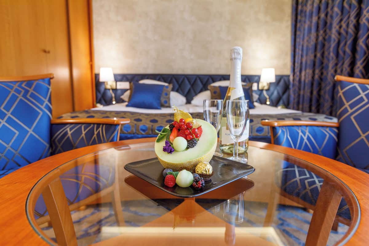 A hotel room with a glass table, a bed with blue patterned pillows, and a decorative fruit arrangement on the table.