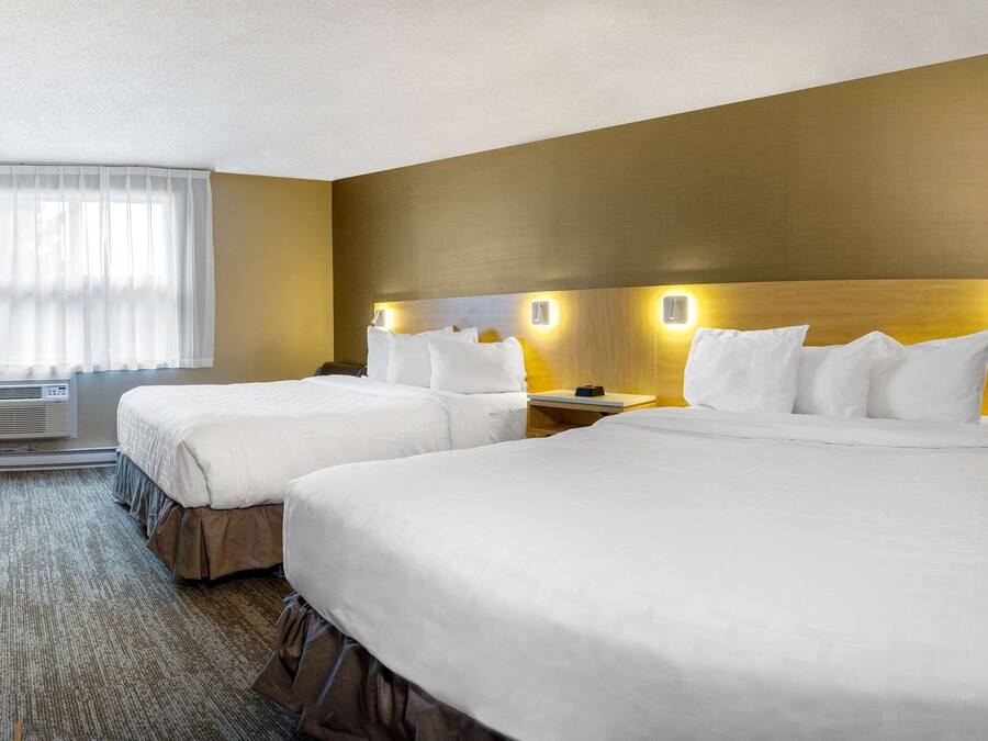 Quarto de hotel com duas camas, uma mesa de cabeceira, uma luminária e uma janela com cortinas.