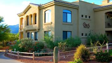 Un edificio con un letrero que dice "The Castas at Sabino Springs". Presenta fachada color crema, varias ventanas y un jardín con cactus y arbustos.