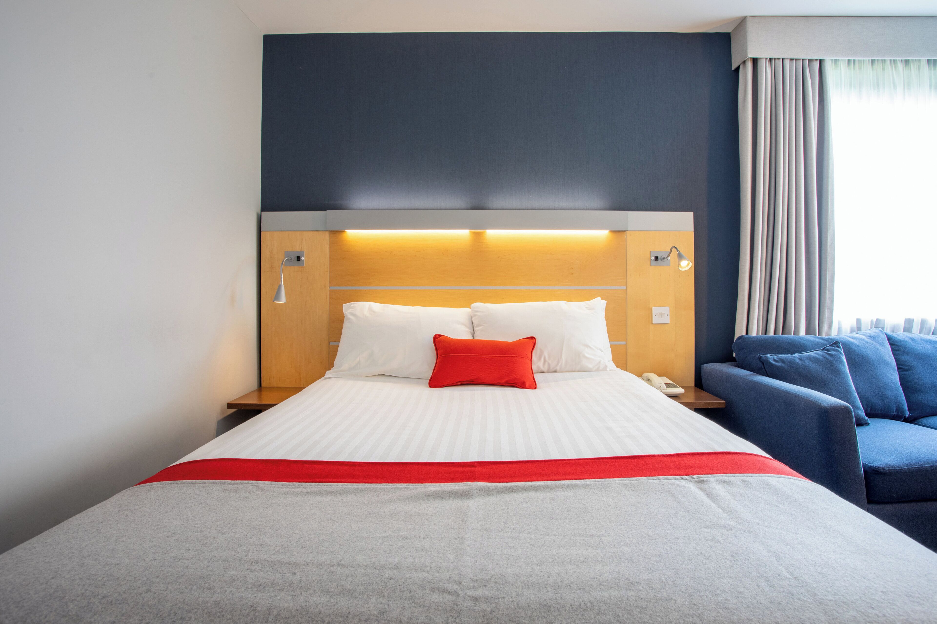 Quarto de hotel com uma cama, mesa de cabeceira com abajur, poltrona azul e uma janela com cortinas.