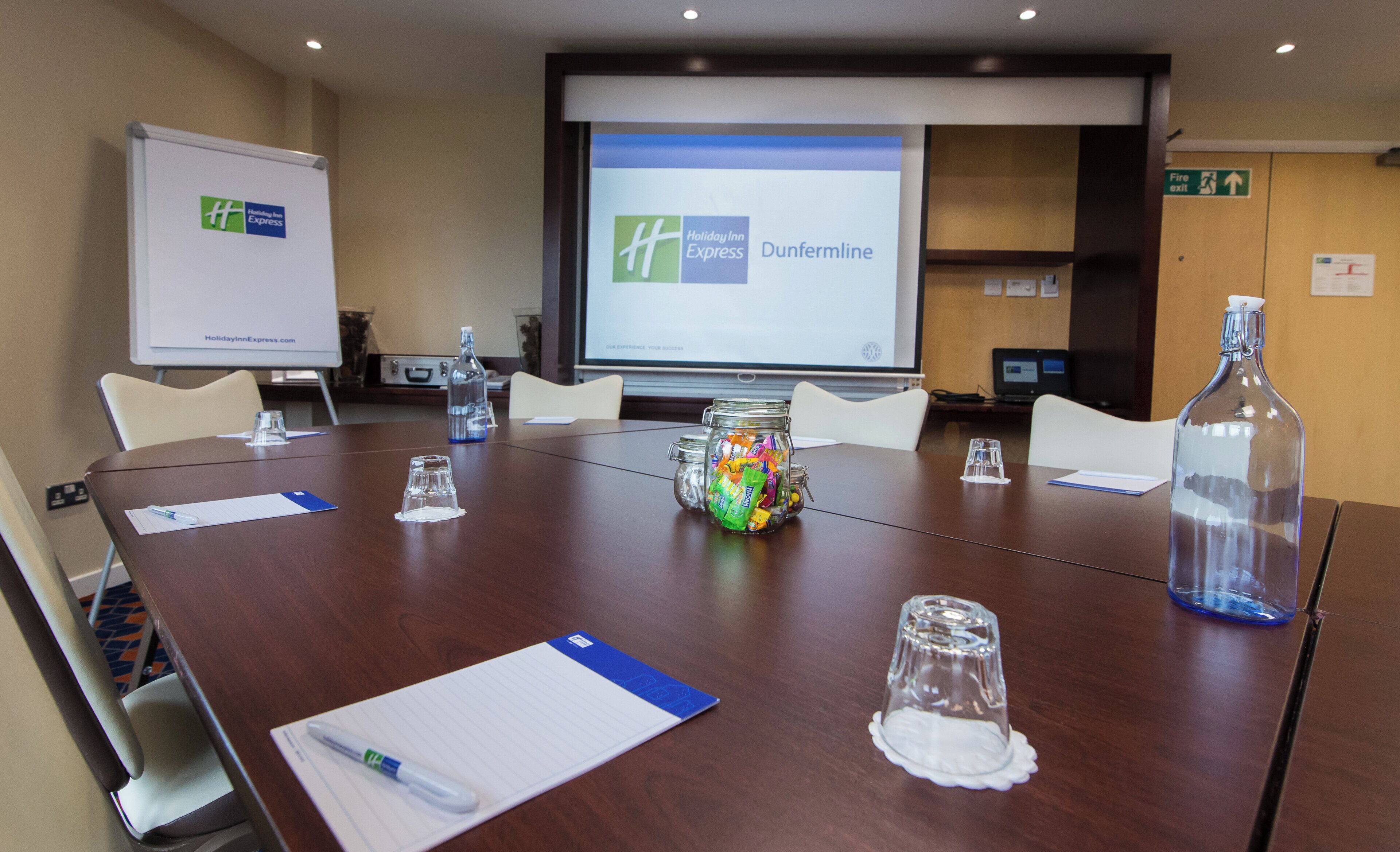 Una sala de conferencias con una mesa grande, sillas, una pantalla de proyector y una pizarra.