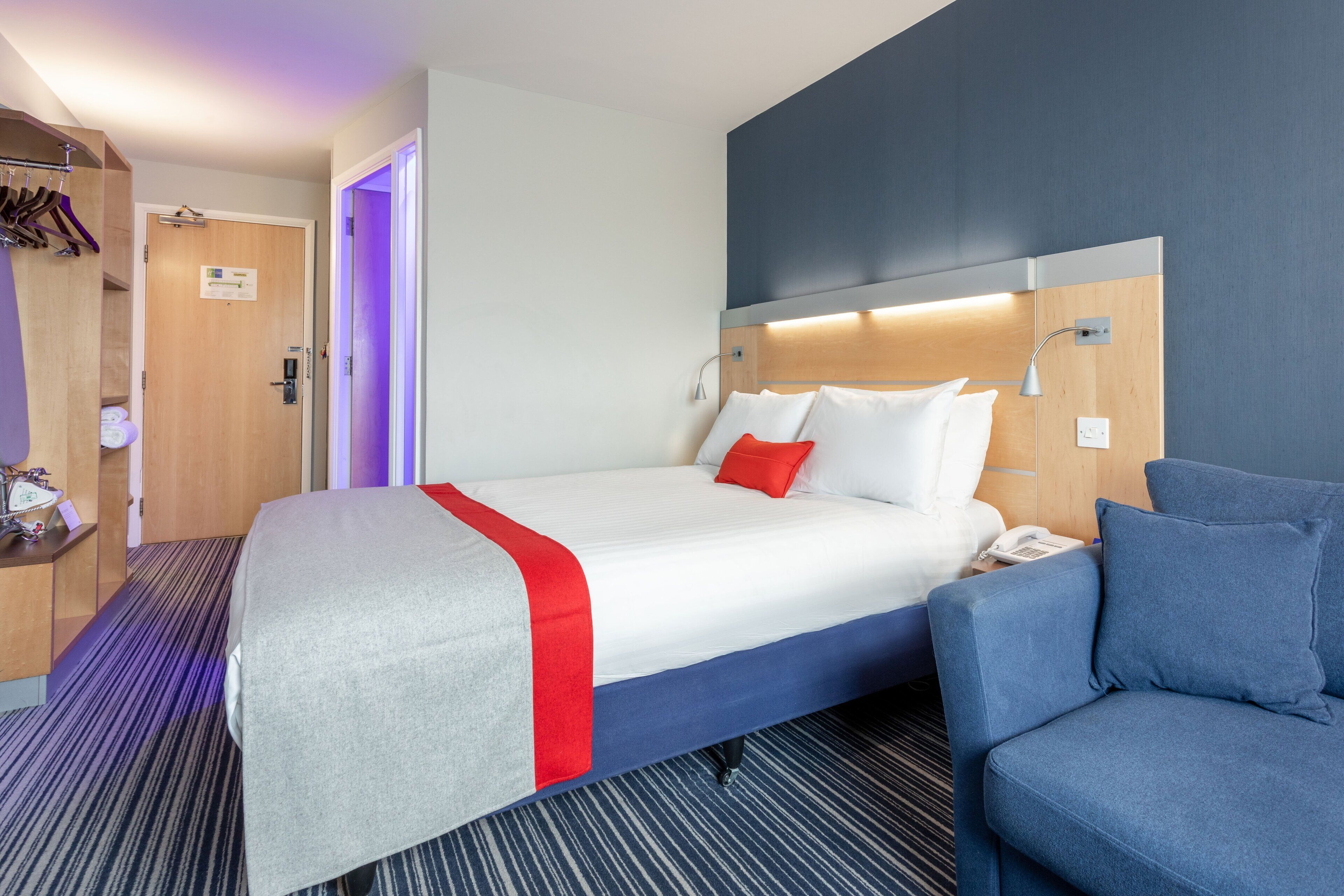 Een hotelkamer met een bed, nachtkastje en een blauwe fauteuil.