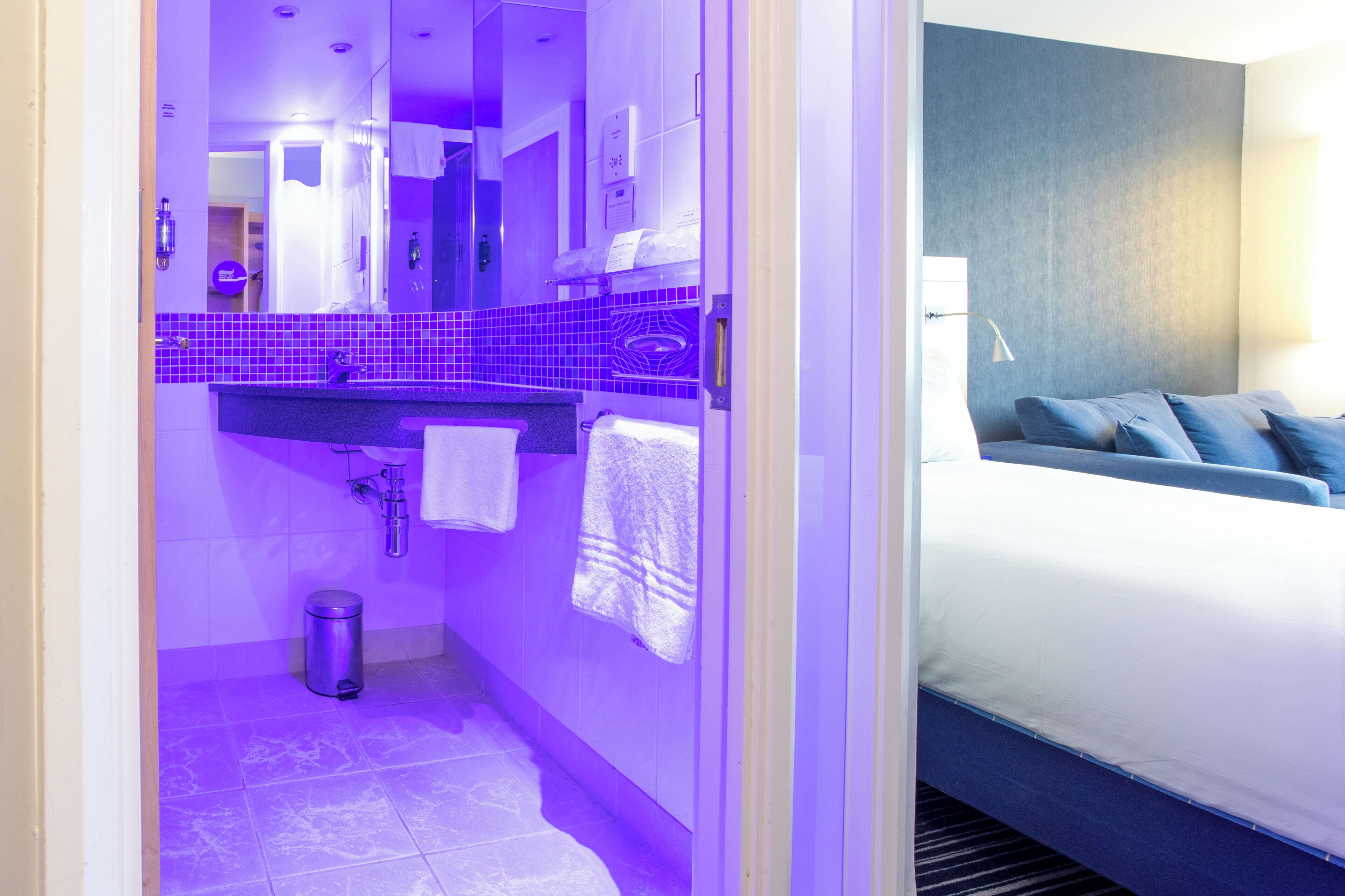 A hotel room with a bathroom featuring purple-tiled walls, a sink, and a mirror. There is a bed with blue bedding and pillows.