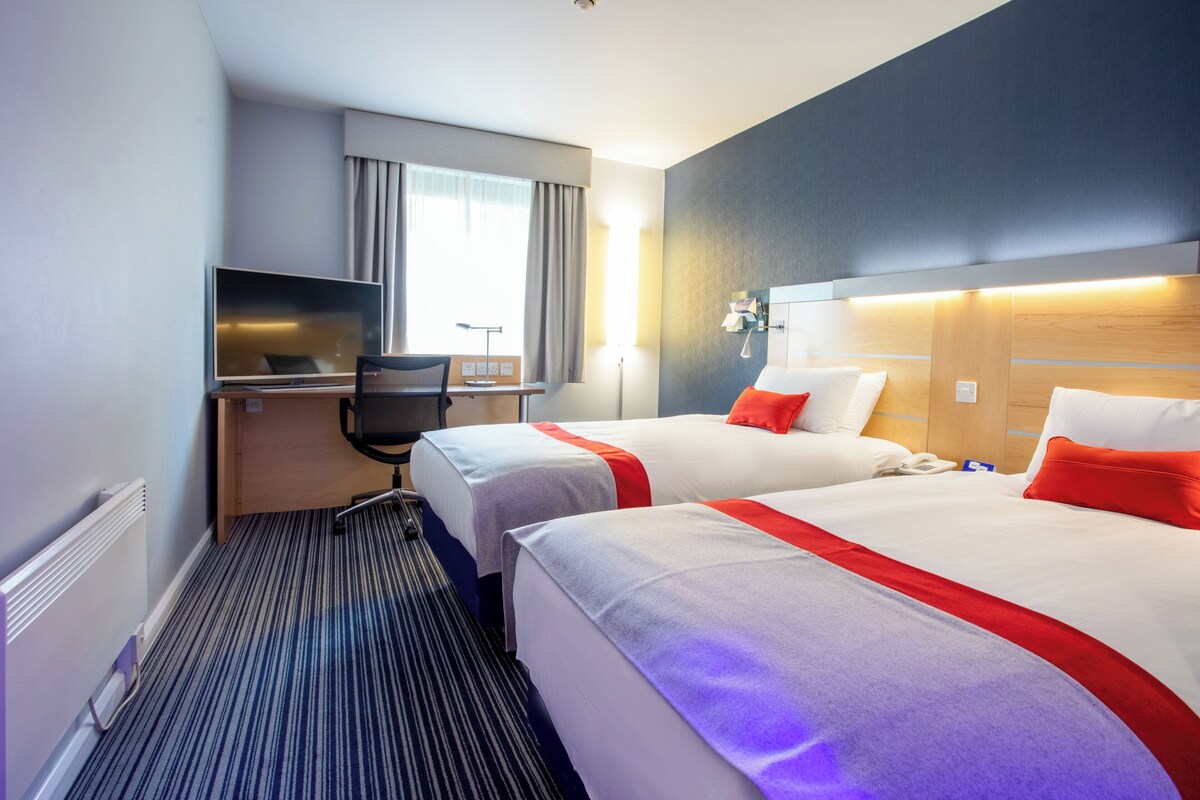 Habitación de hotel con dos camas, un escritorio con silla, televisor y ventana con cortinas.
