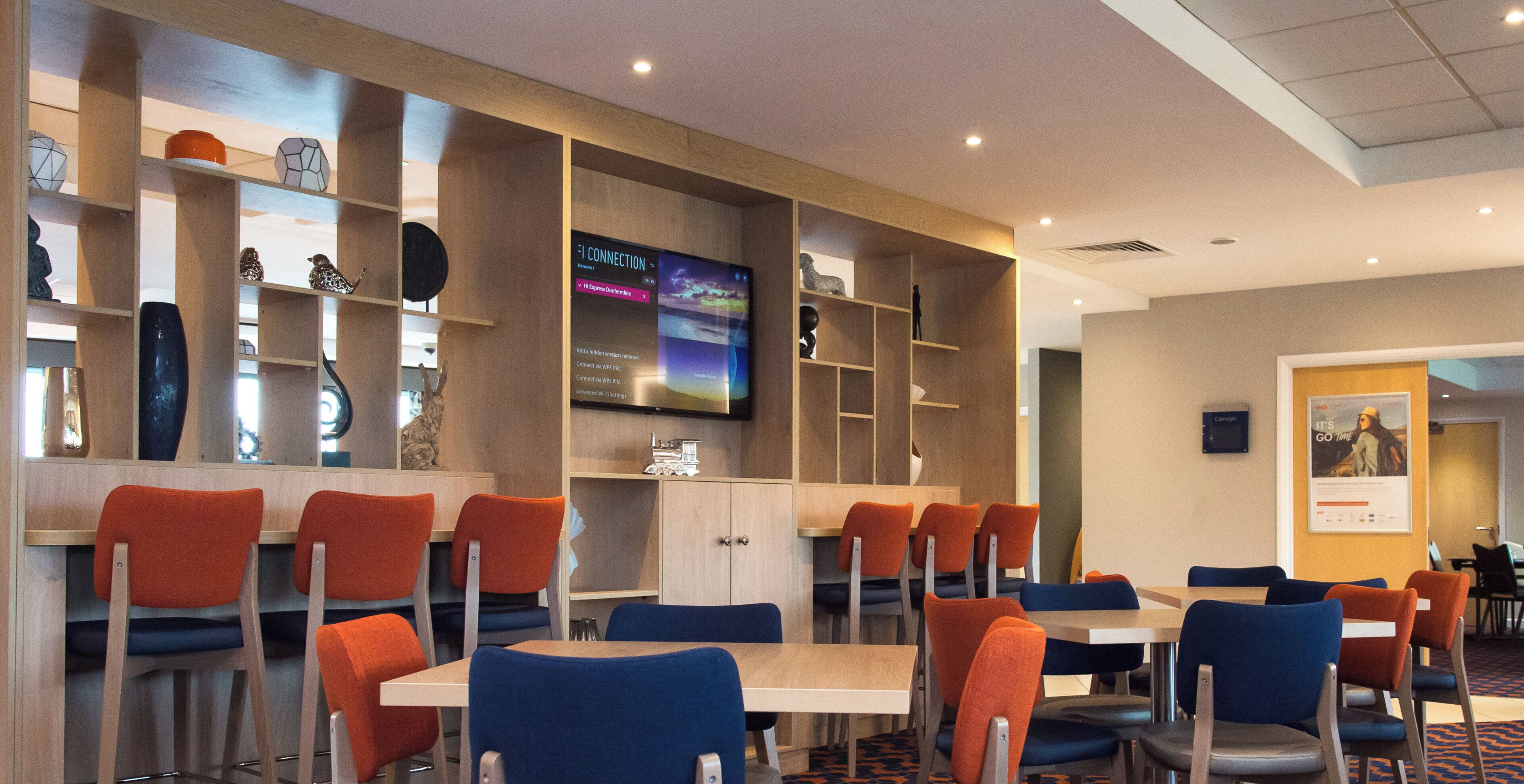 Habitación de hotel con televisor montado en la pared, una estantería con objetos decorativos y una zona de estar con sillas y mesas.