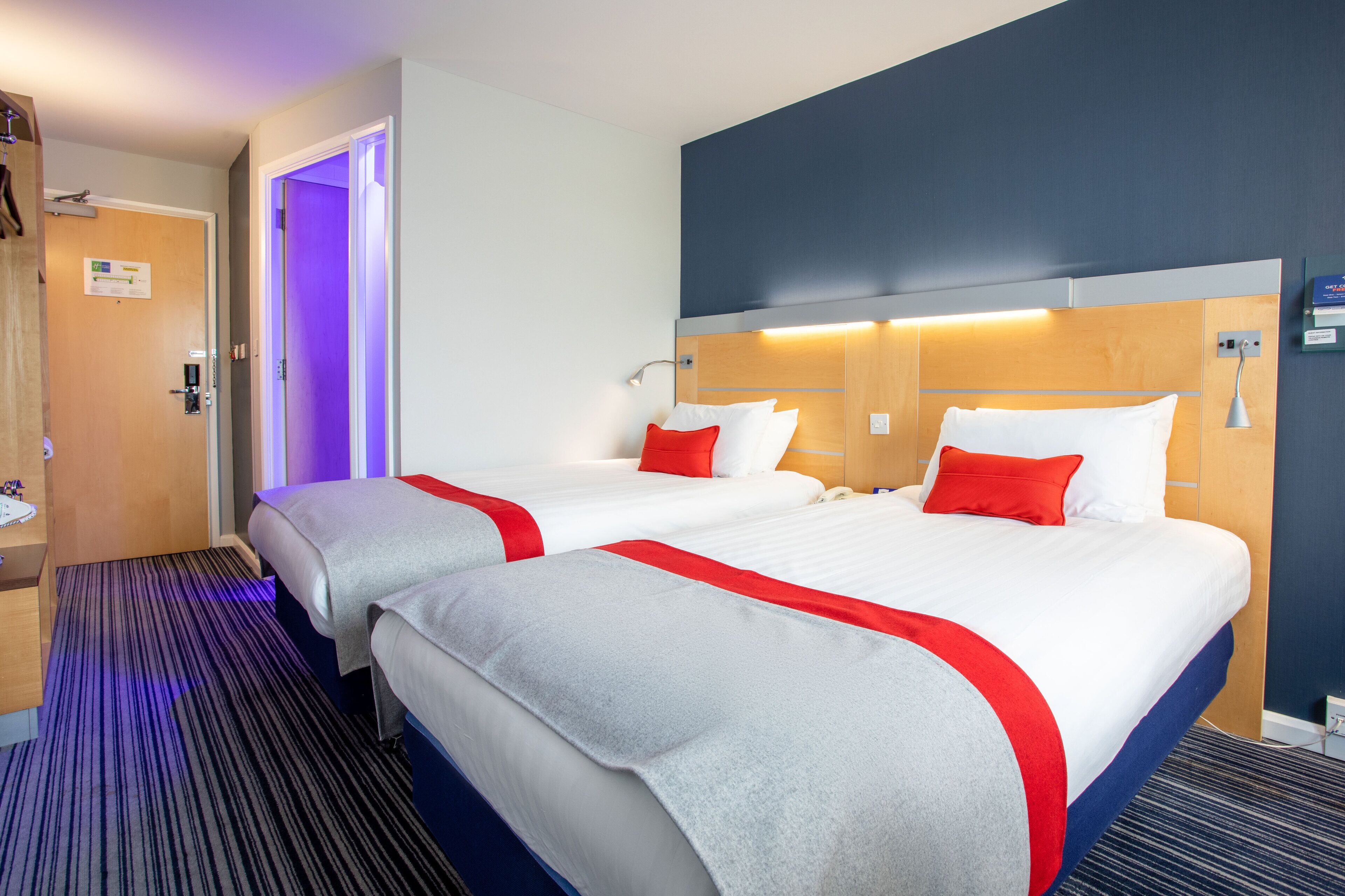 Hotelkamer met twee bedden, elk met witte beddengoed en rode accenten, een houten hoofdeinde, een nachtkastje en een gestreept tapijt.