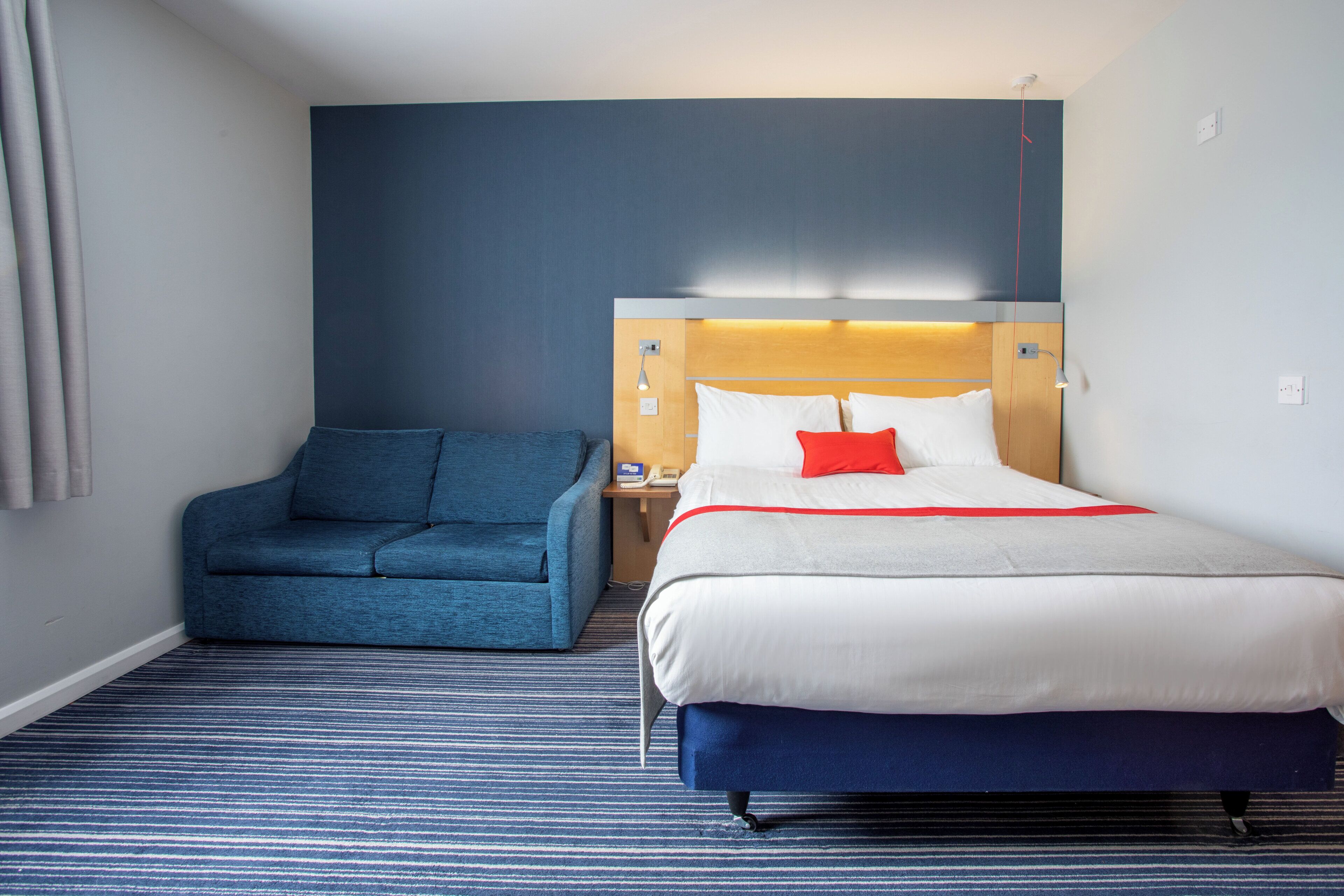 A hotel room with a bed, a blue sofa, a wooden headboard, and a striped carpet.