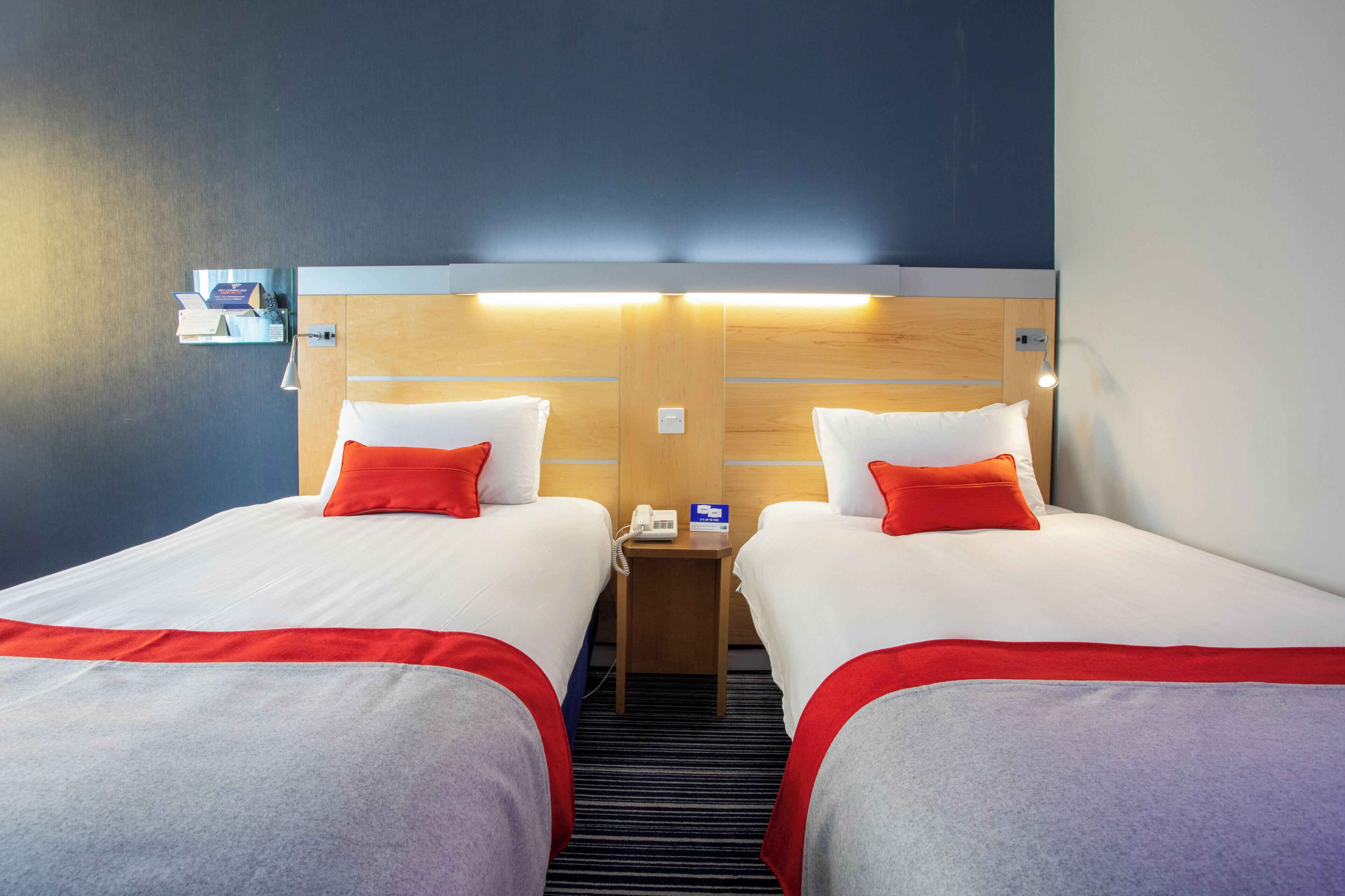 Habitación de hotel con dos camas, cada una con ropa blanca y detalles rojos, un cabecero de madera, una mesita de noche con teléfono, y una lámpara de pared.