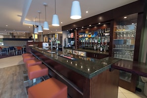 Uma área de bar moderna com um balcão comprido, diversas torneiras e seleção de garrafas e copos.
