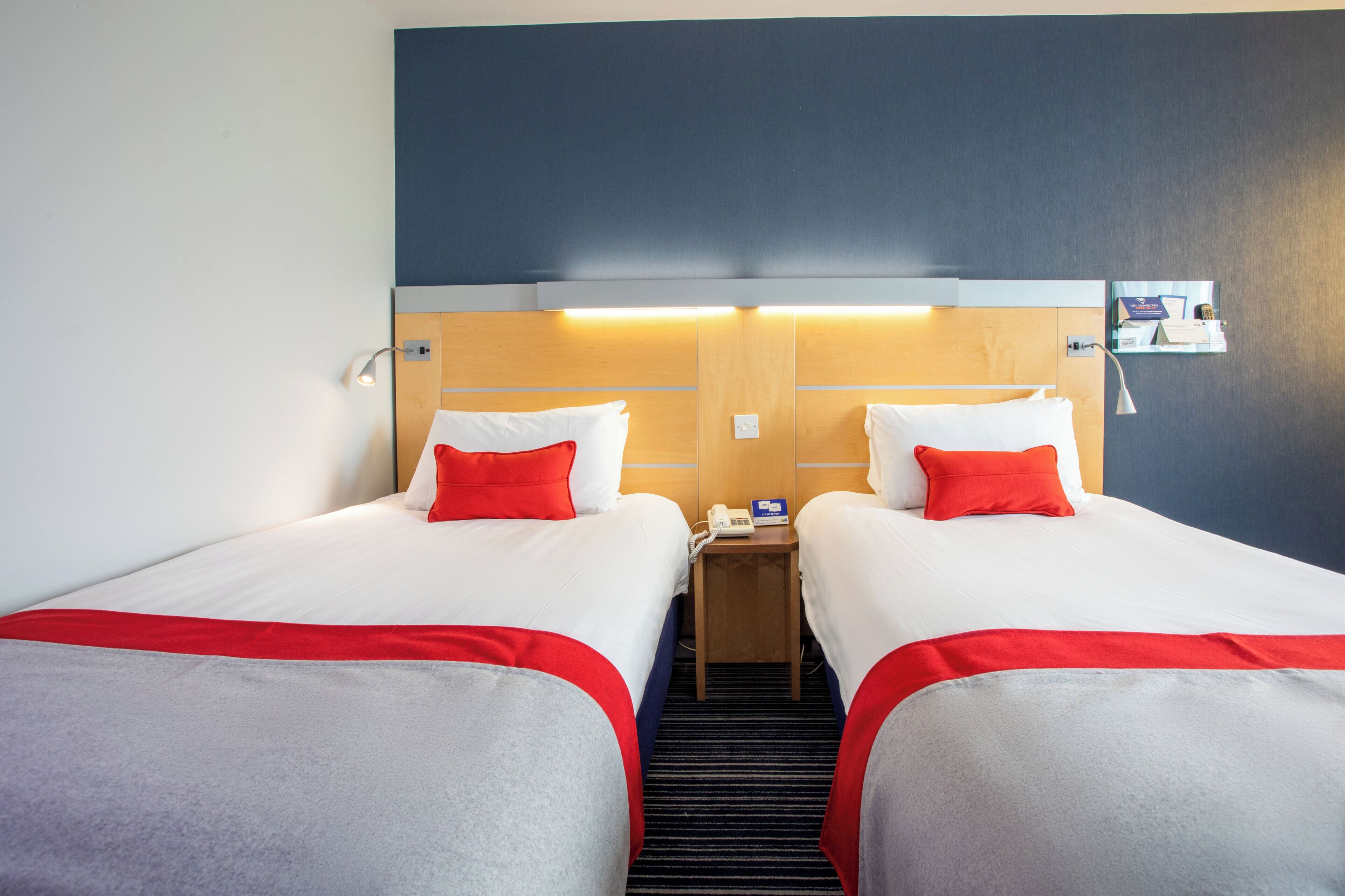 A hotel room with two beds, each with white bedding and red accents, a bedside table with a phone and a card holder, and a wall-mounted light fixture.