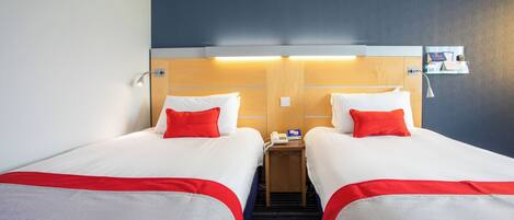 A hotel room with two beds, each with white bedding and red accents, a bedside table with a phone and a card holder, and a wall-mounted light fixture.