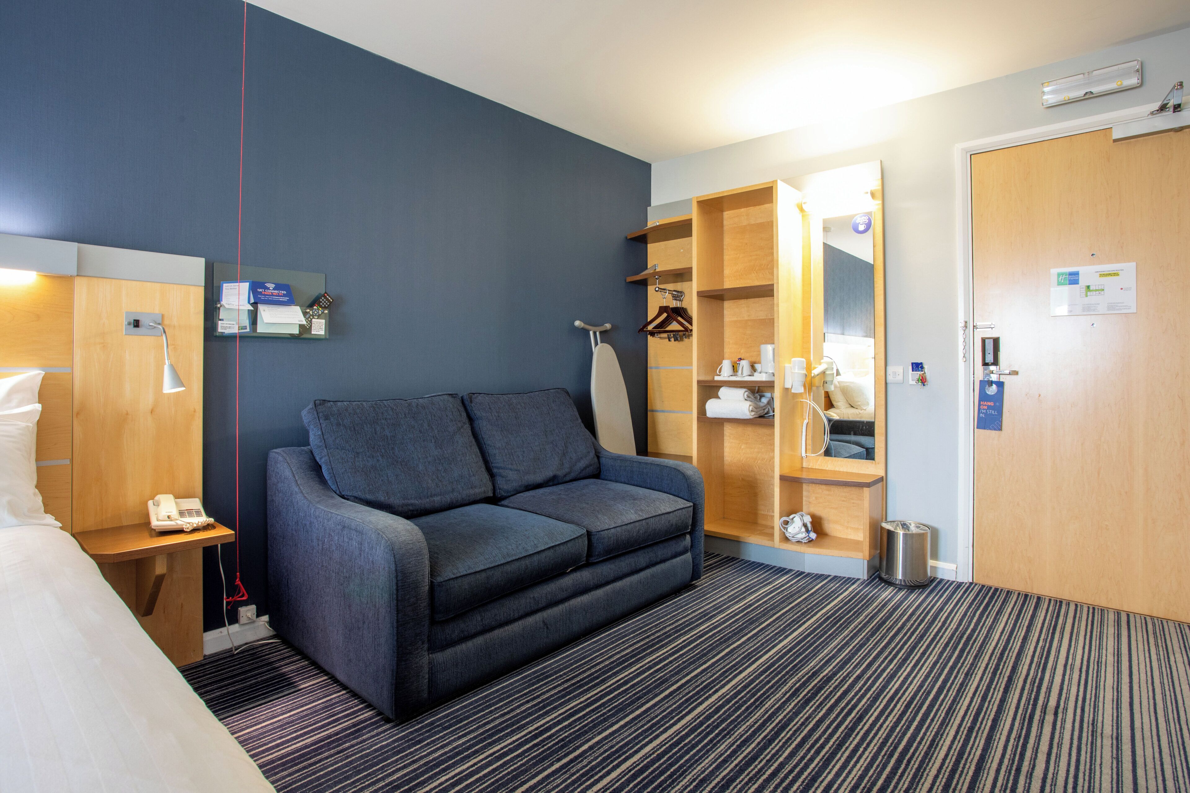A hotel room with a sofa, a desk with a lamp, a wardrobe, and a door.