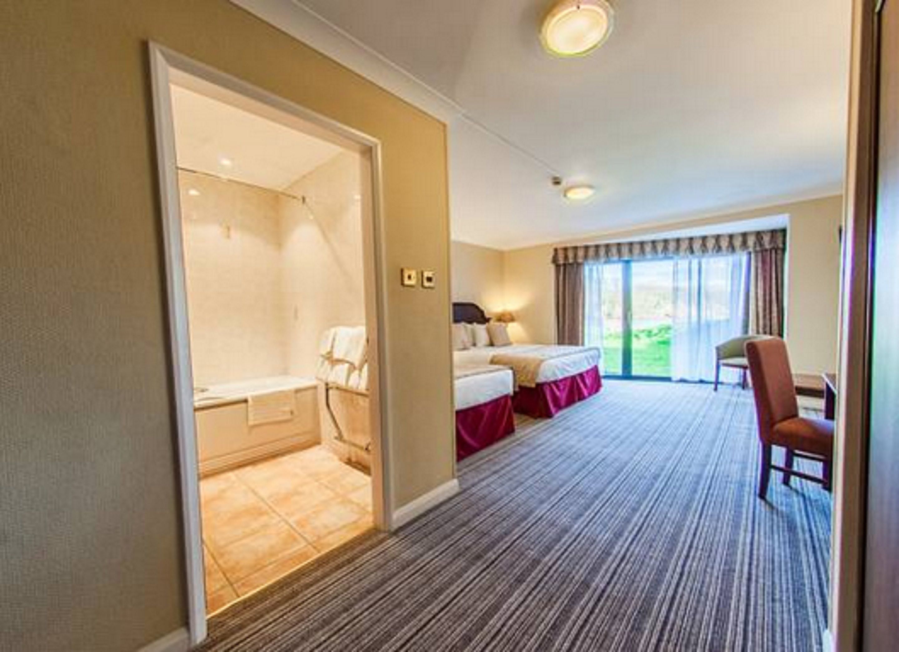 A hotel room with a large bed, a separate bathroom, a seating area with a chair, and a view of the outdoors through a sliding door.