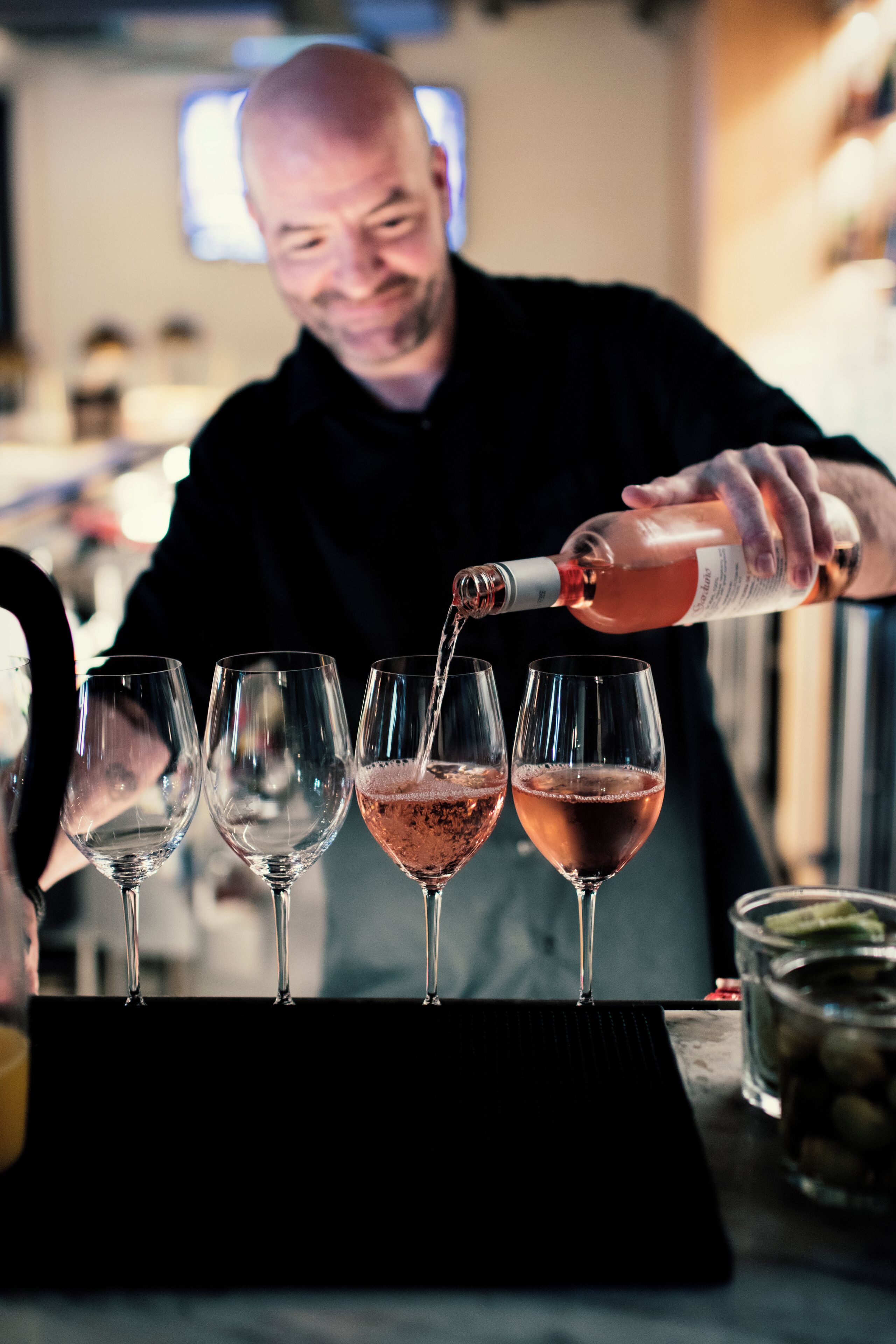 Une personne verse un liquide rose dans quatre verres sur le comptoir d’un bar.