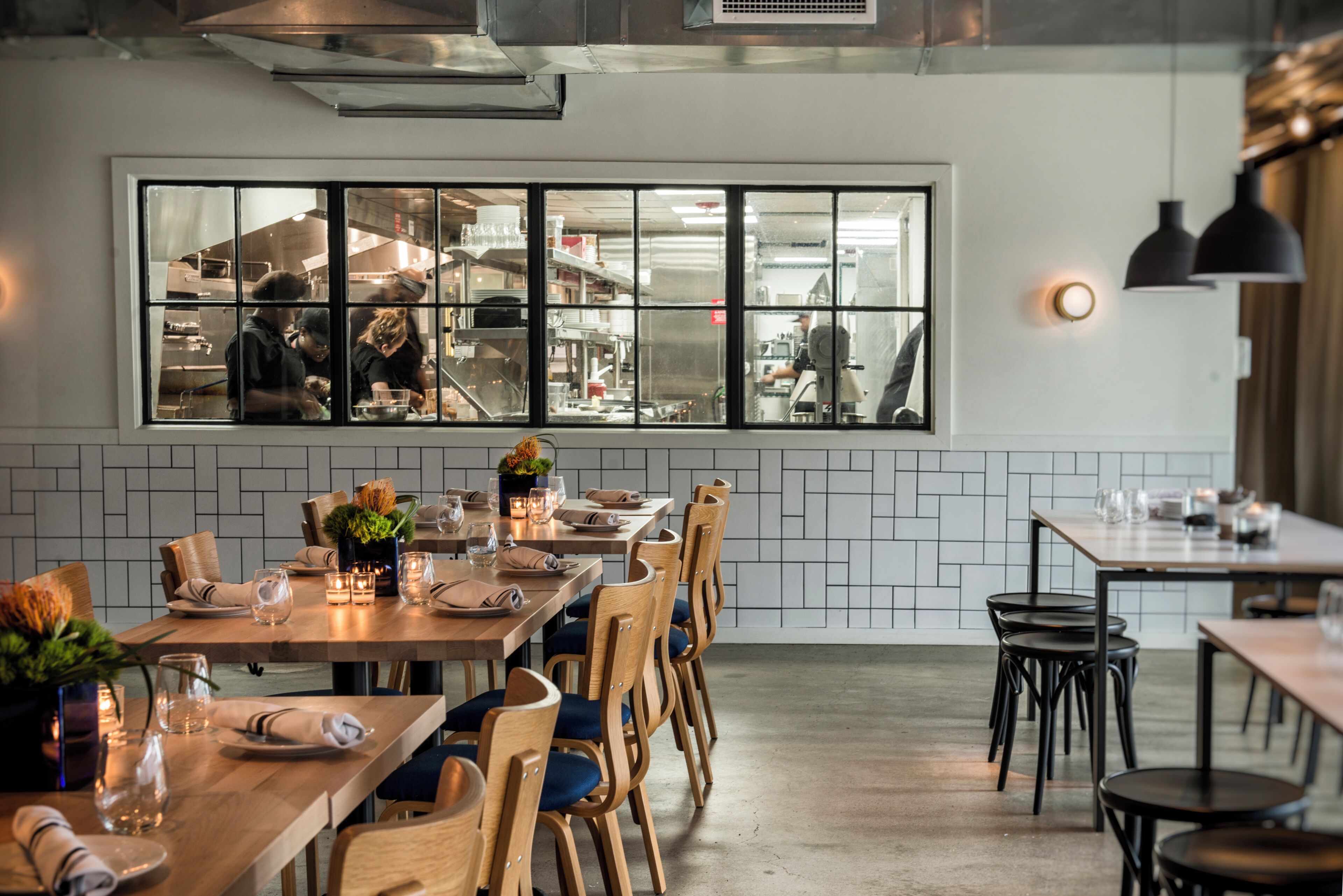 Ein modernes Restaurant-Interieur mit einem großen Fenster zum Blick auf die Küche, Holztische mit Stühlen und einem sauberen, gefliesten Boden.