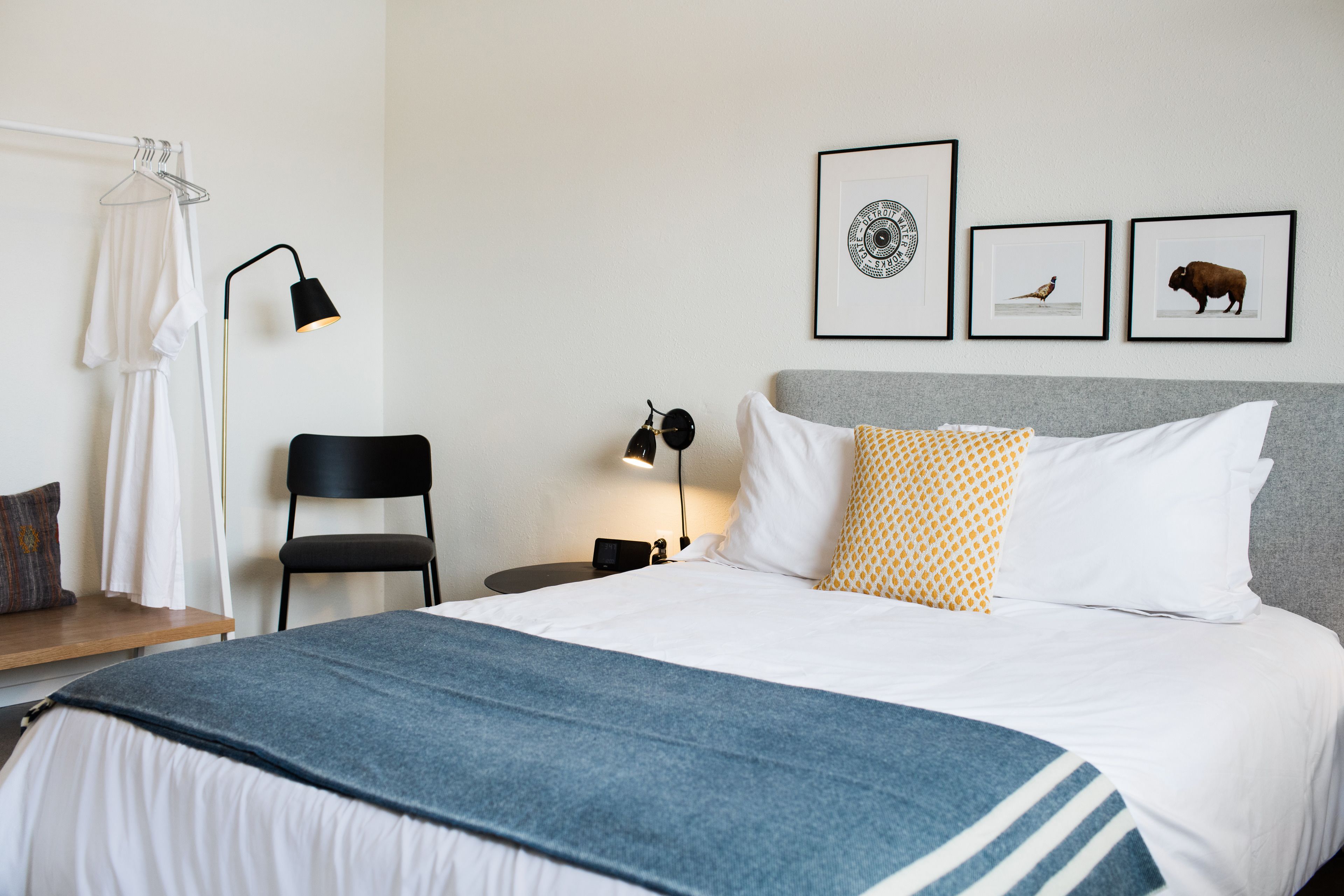 A neatly arranged hotel room with a bed, a chair, a bedside table with a lamp, and framed pictures on the wall.