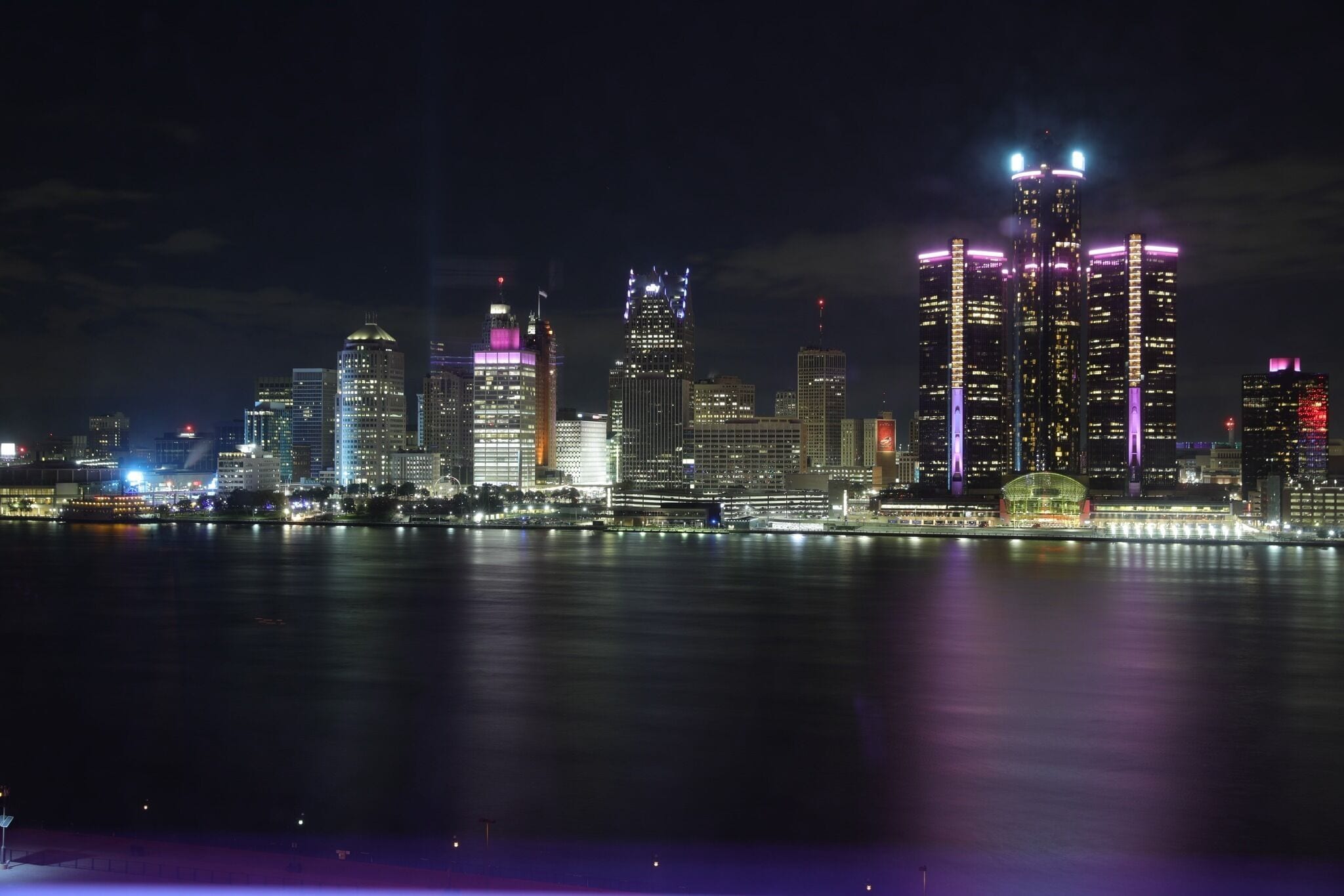 A nighttime city skyline with illuminated buildings along a waterfront.