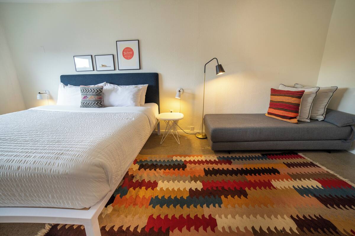 A bedroom with a bed, a sofa, a patterned rug, and wall-mounted lights.