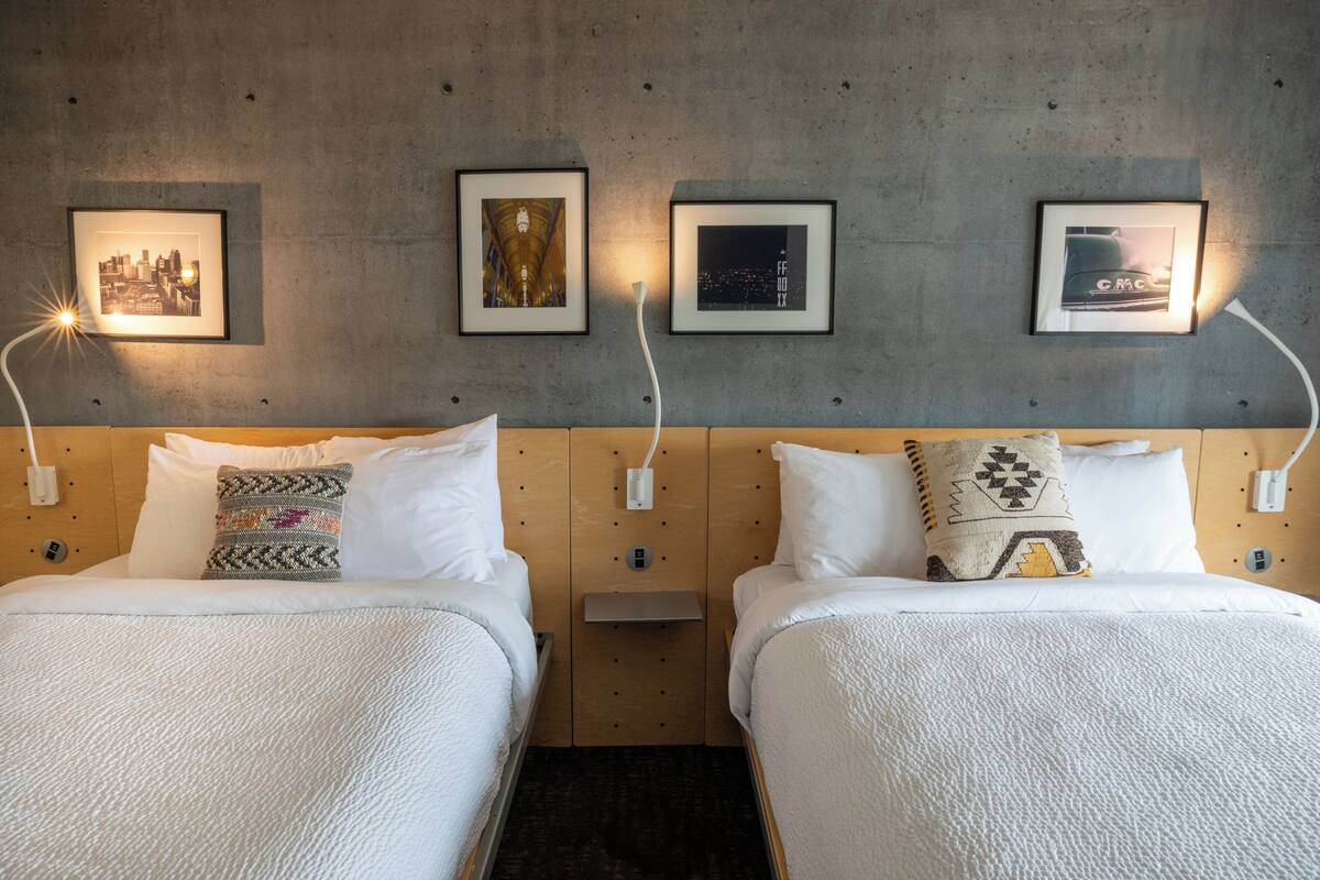 A hotel room with two beds, each with white bedding and decorative pillows. There are framed pictures on the wall above the beds, and a small shelf between the beds.
