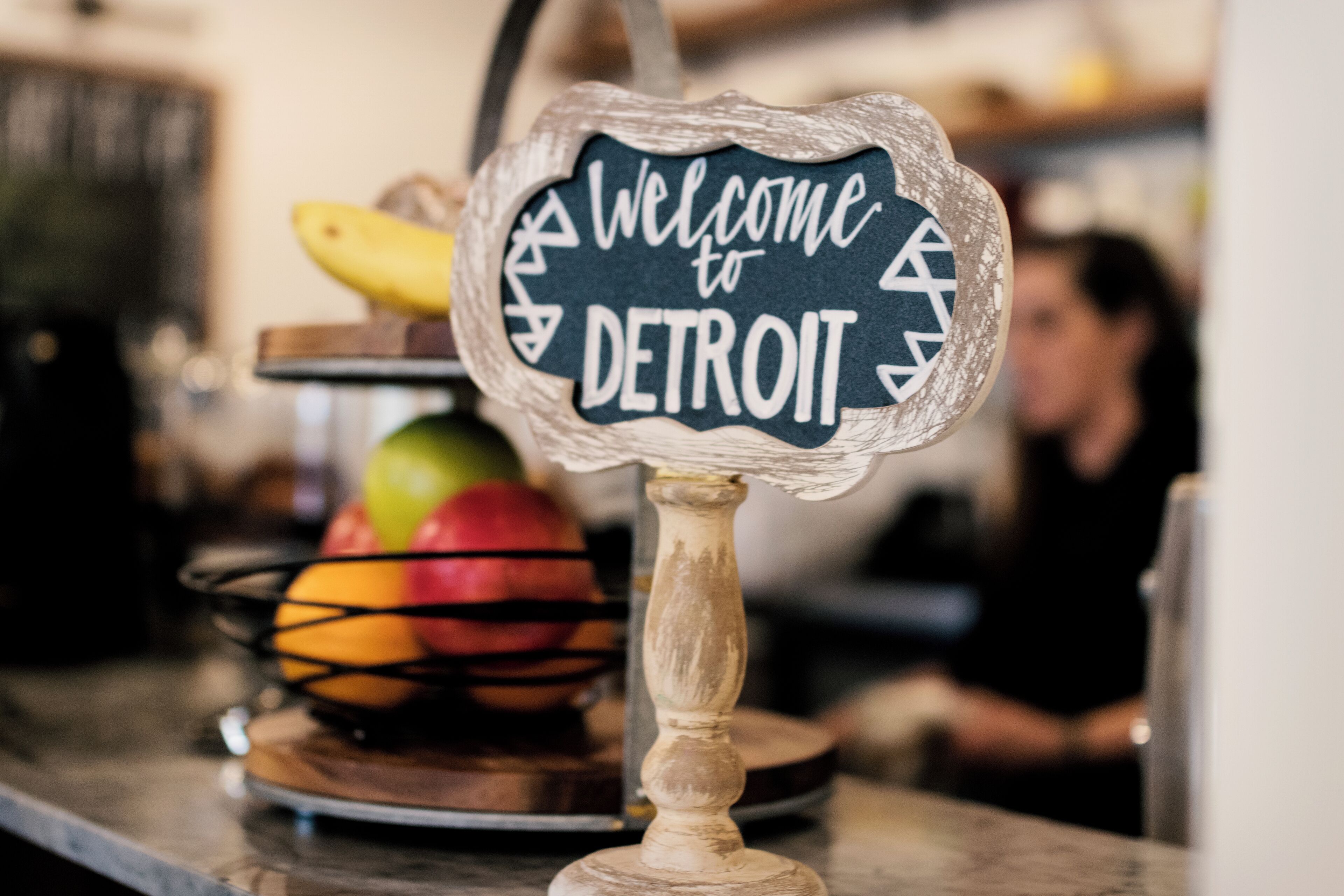 A welcome sign to Detroit on a rustic wooden stand with a fruit bowl behind it.