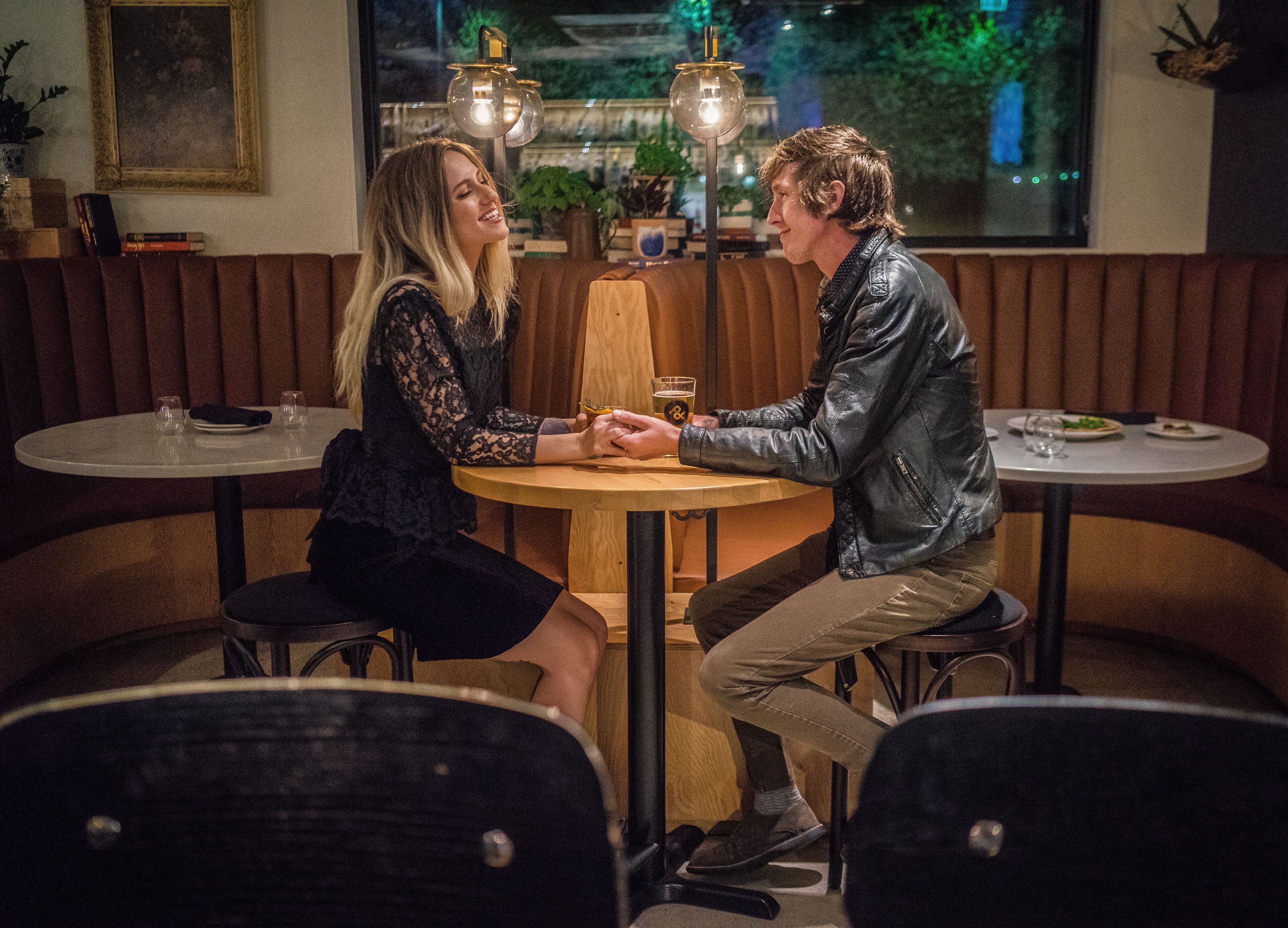 A couple seated at a round table in a restaurant with wooden booths and pendant lighting.