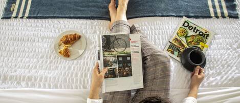 Uma pessoa deitada na cama com uma revista, um croissant e uma chávena.