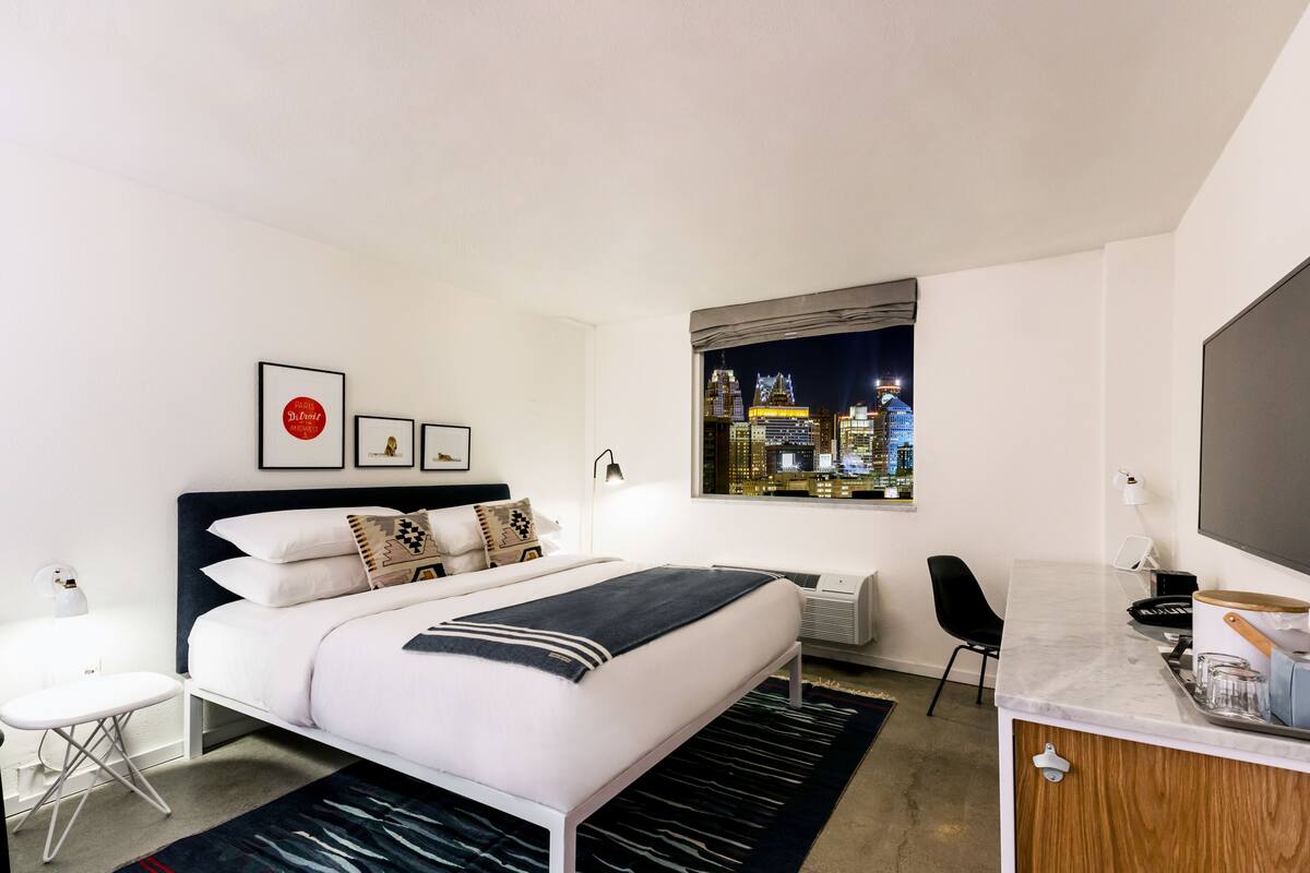 A modern hotel room with a bed, a desk, a chair, a window with a city view, and a flat-screen TV.