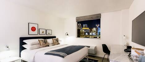 Ein modernes Hotelzimmer mit einem Bett, einem Schreibtisch, einem Stuhl, einem Fenster mit Blick auf die Stadt und einem Flachbildfernseher.
