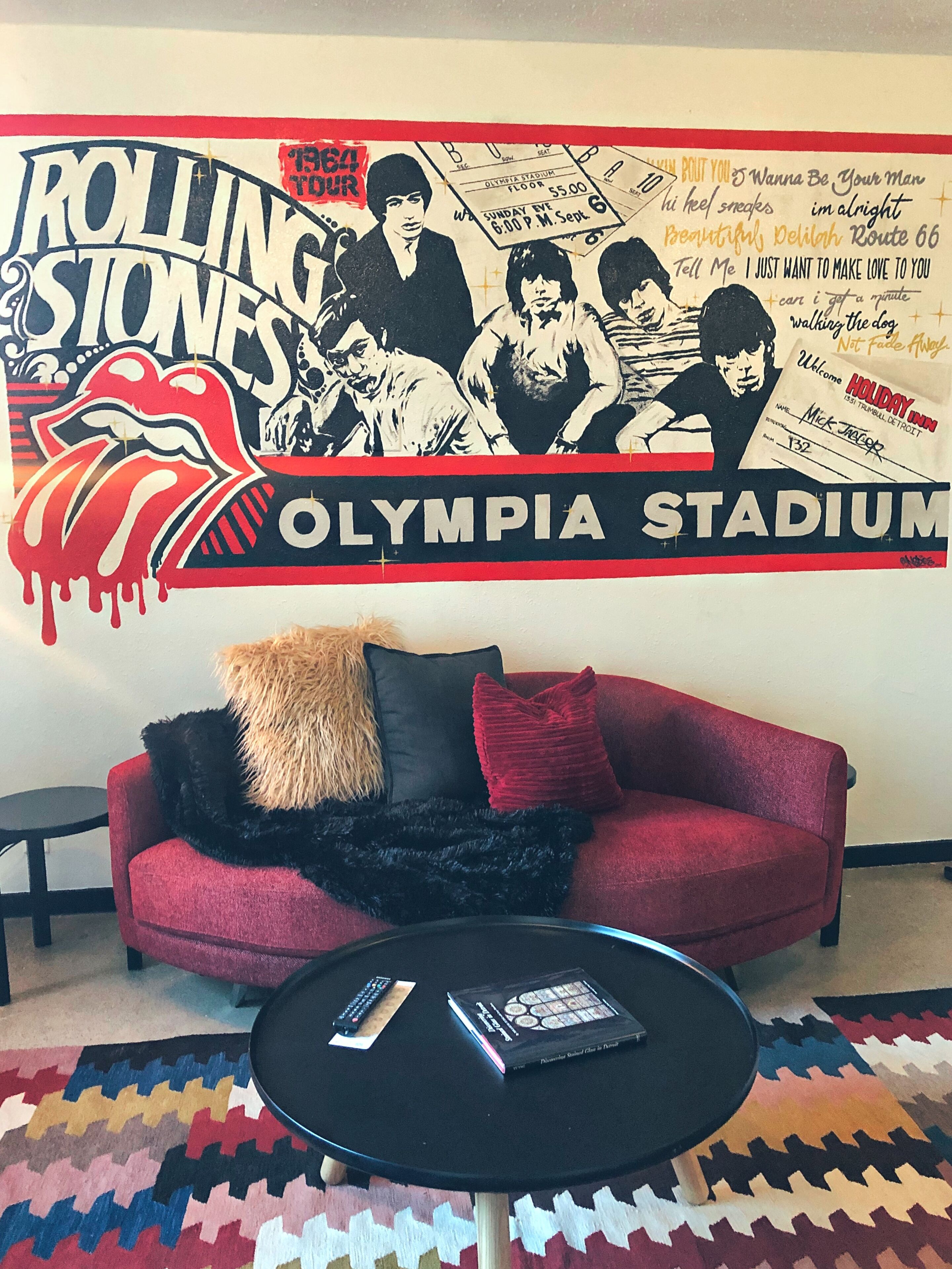 Una habitación con un sofá rojo, almohadas negras y rojas, una mesa de centro negra con un control remoto y un libro, y una alfombra colorida.