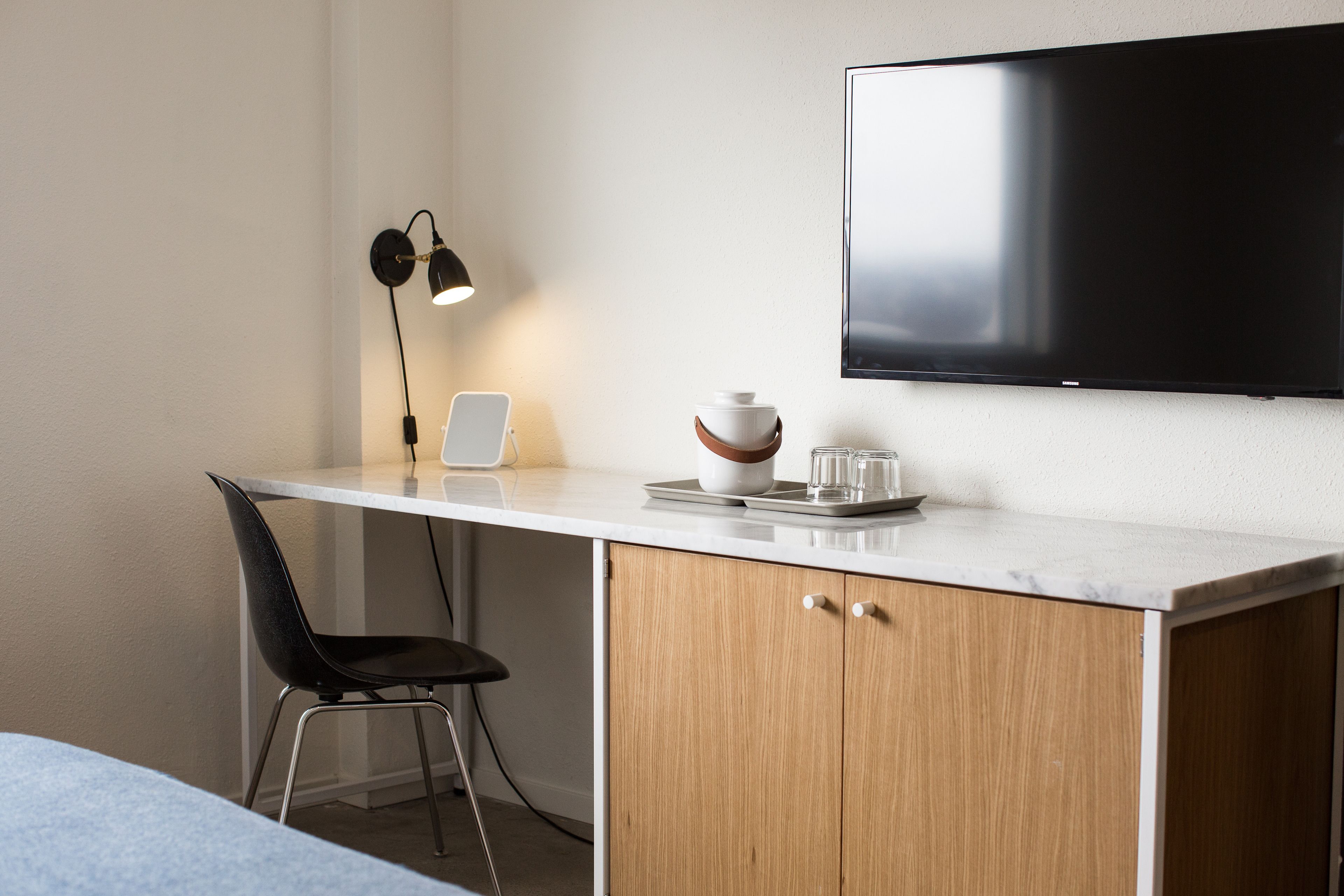 A modern hotel room with a white desk, a black chair, a flat-screen TV mounted on the wall, a small table with a bowl and glasses, and a desk lamp.