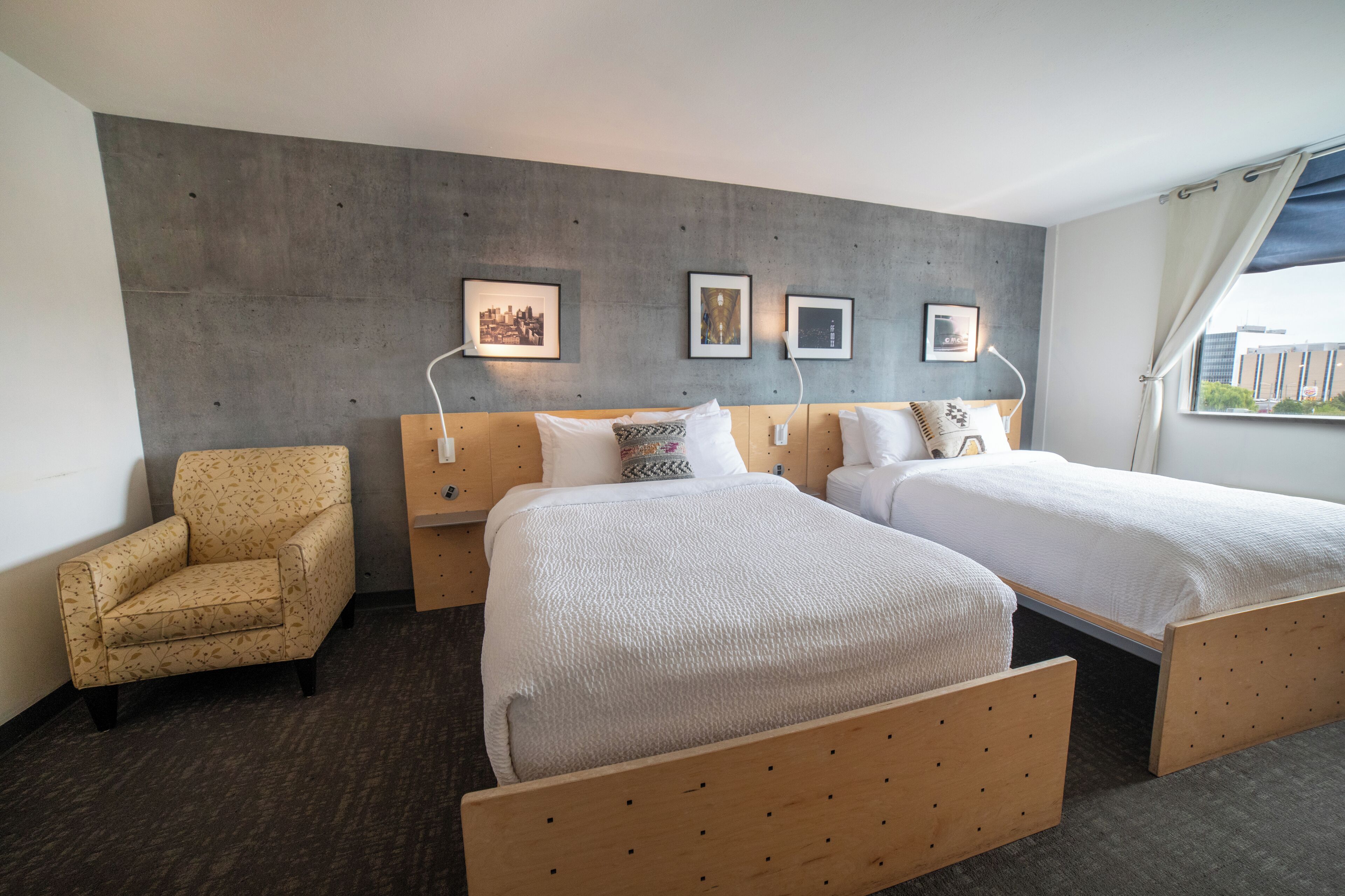 A hotel room with two beds, a chair, and a concrete accent wall with framed pictures.